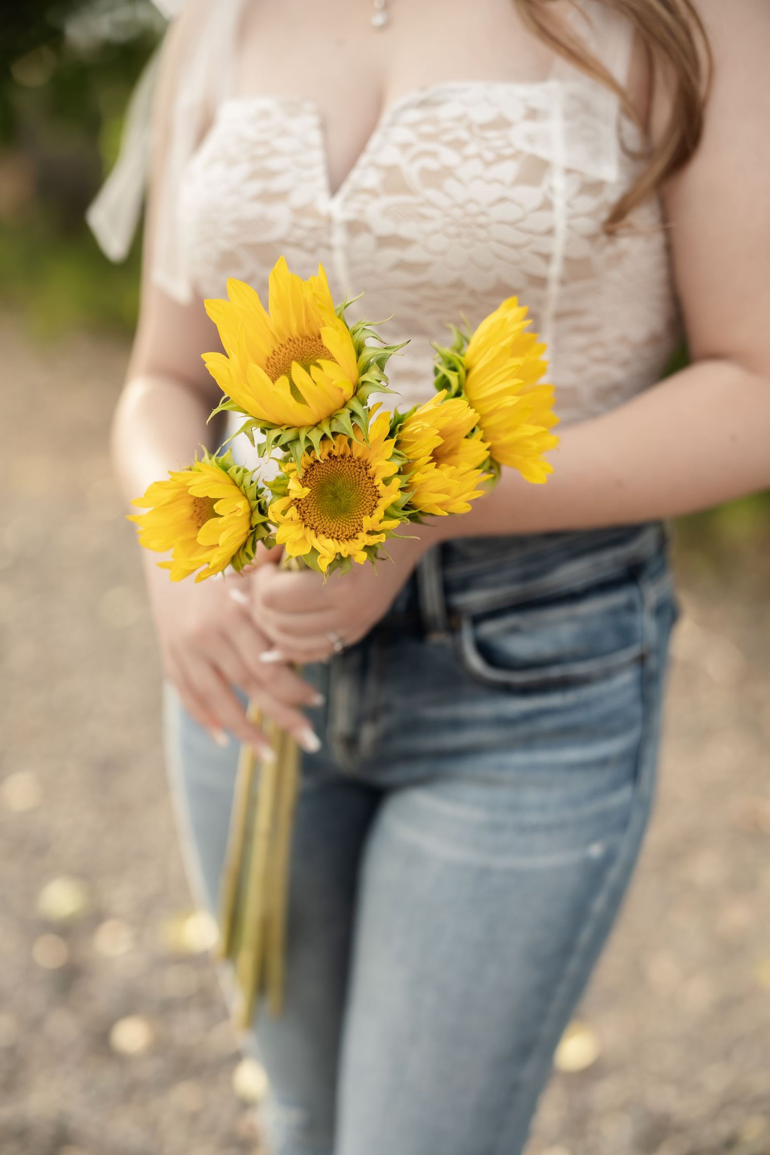 Someone holding vibrant yellow sunflowers against a white lace top and denim jeans outdoors.