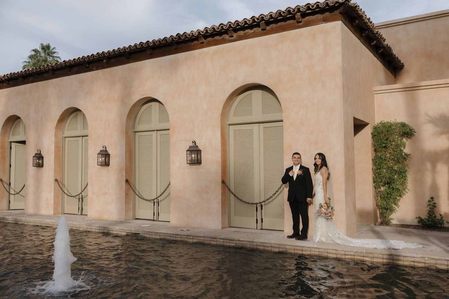 Bride and groom pose by a fountain outside a Spanish-style venue with arched doors and warm stucco walls.