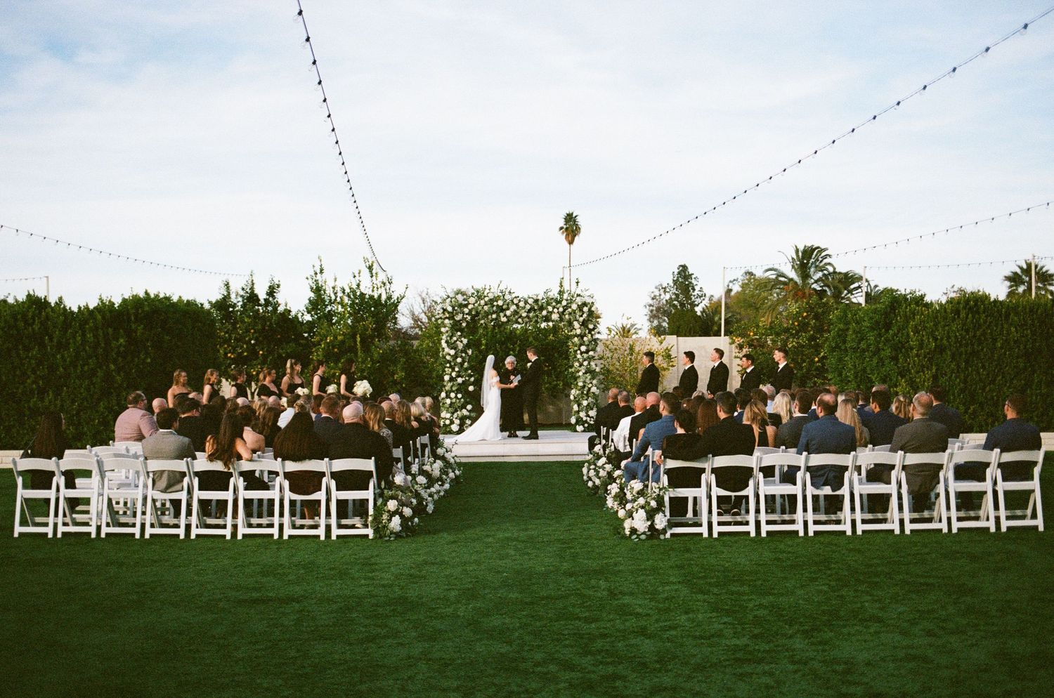 Outdoor wedding ceremony on lush green lawn with white chairs, floral arch altar, string lights, and lush hedges backdrop.