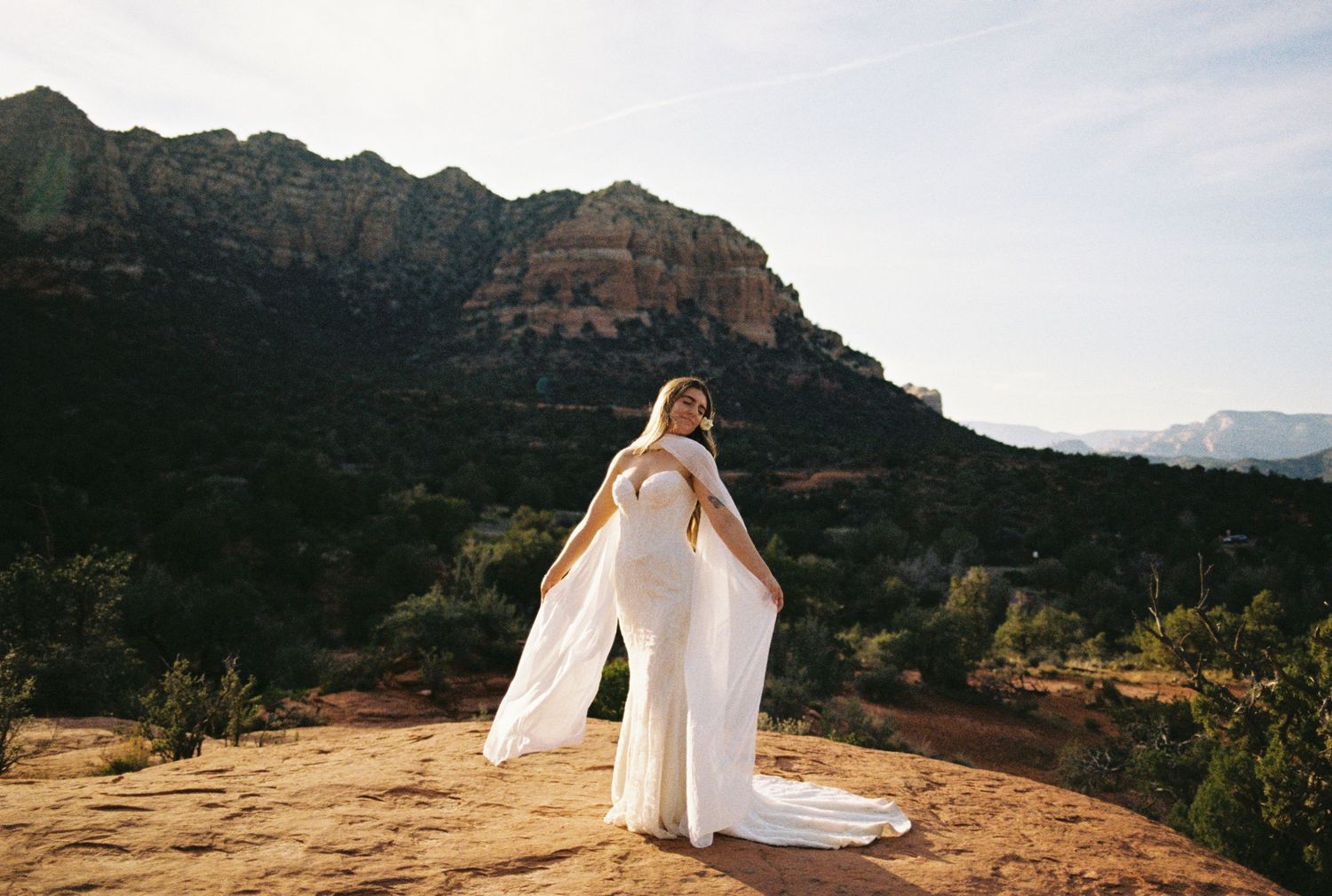 Bride in white gown with flowing cape poses on red rock in Sedona, Arizona, with stunning desert landscape backdrop.