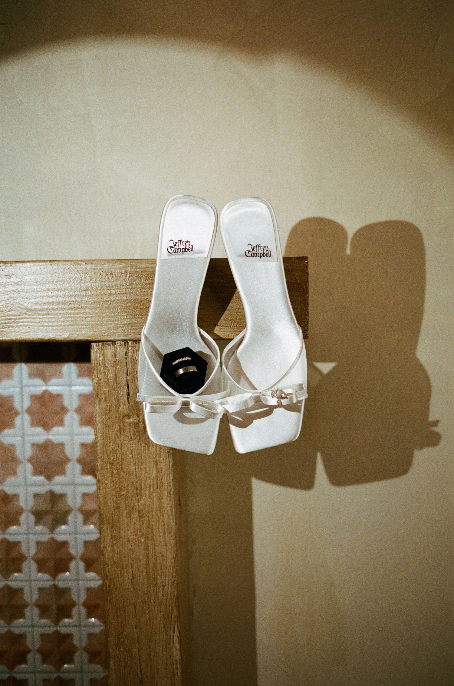 White bridal heels with a wedding ring resting inside, casting elegant shadows on a beige wall.