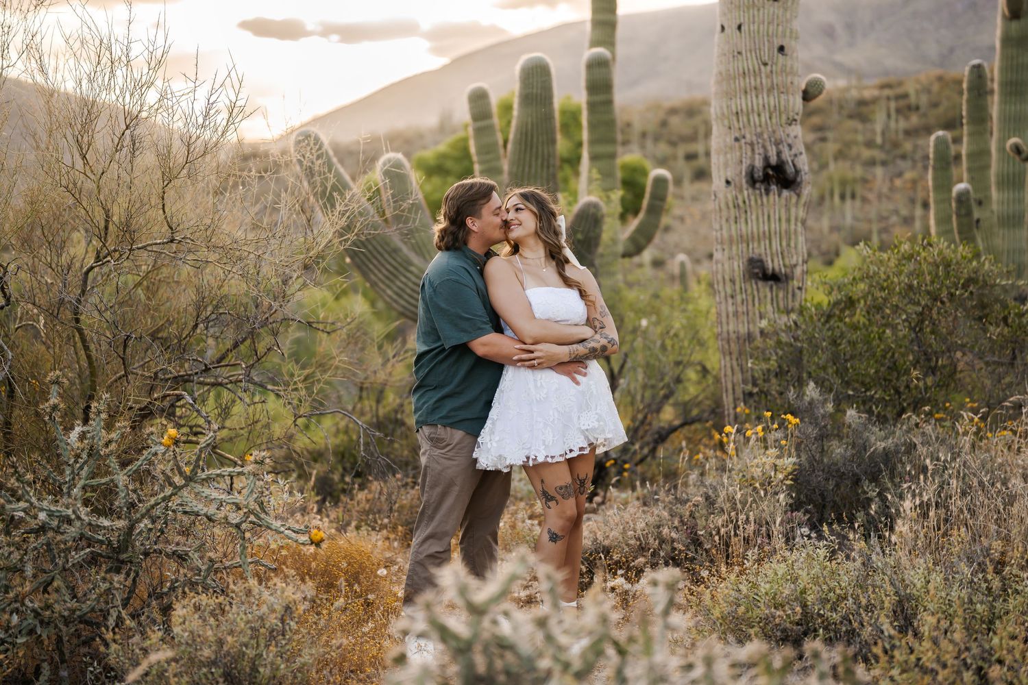 A romantic couple embraces in front of blooming saguaro cacti and desert landscape during golden hour in Arizona.