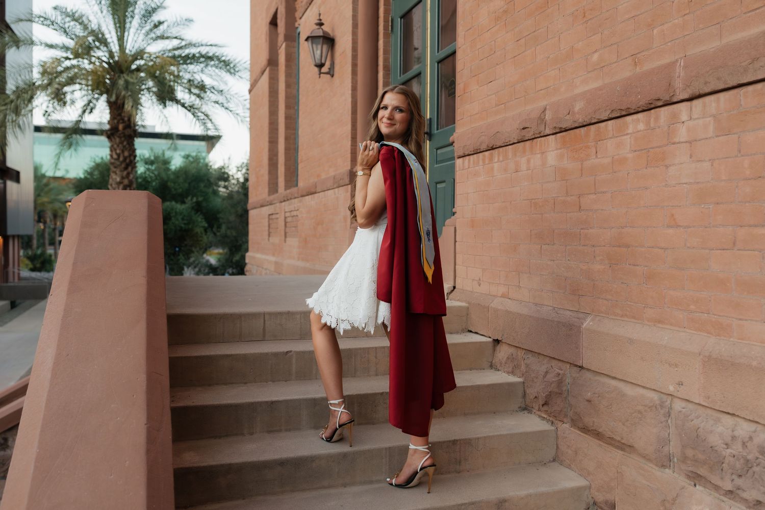 Woman in white feathered dress with red cape poses on stone steps beside a terracotta building with palm tree.