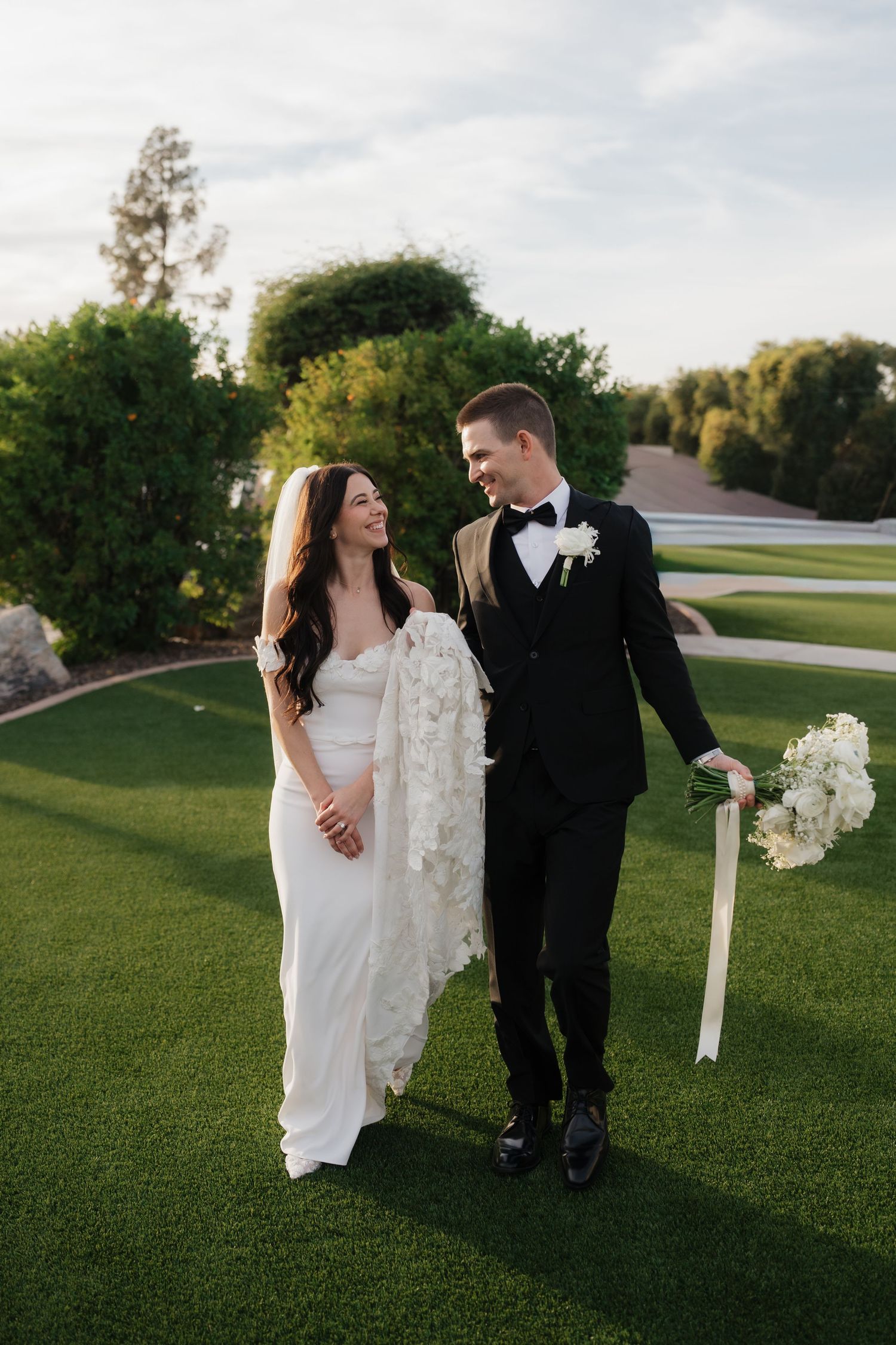 Newlyweds walking together on manicured lawn during golden hour with white floral bouquet.