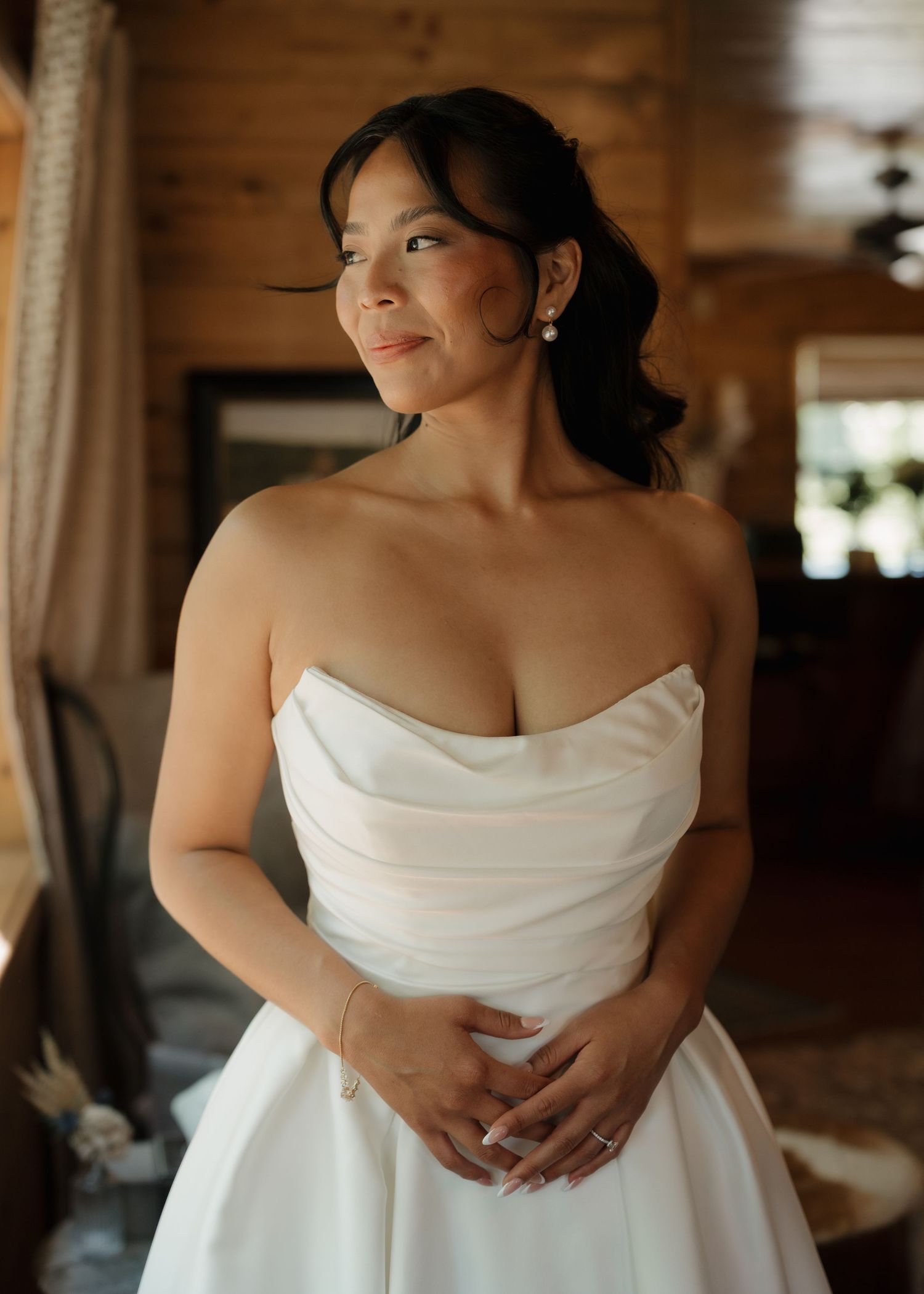 Bride in strapless white satin wedding gown smiling softly, hands clasped, in a rustic wooden cabin setting.