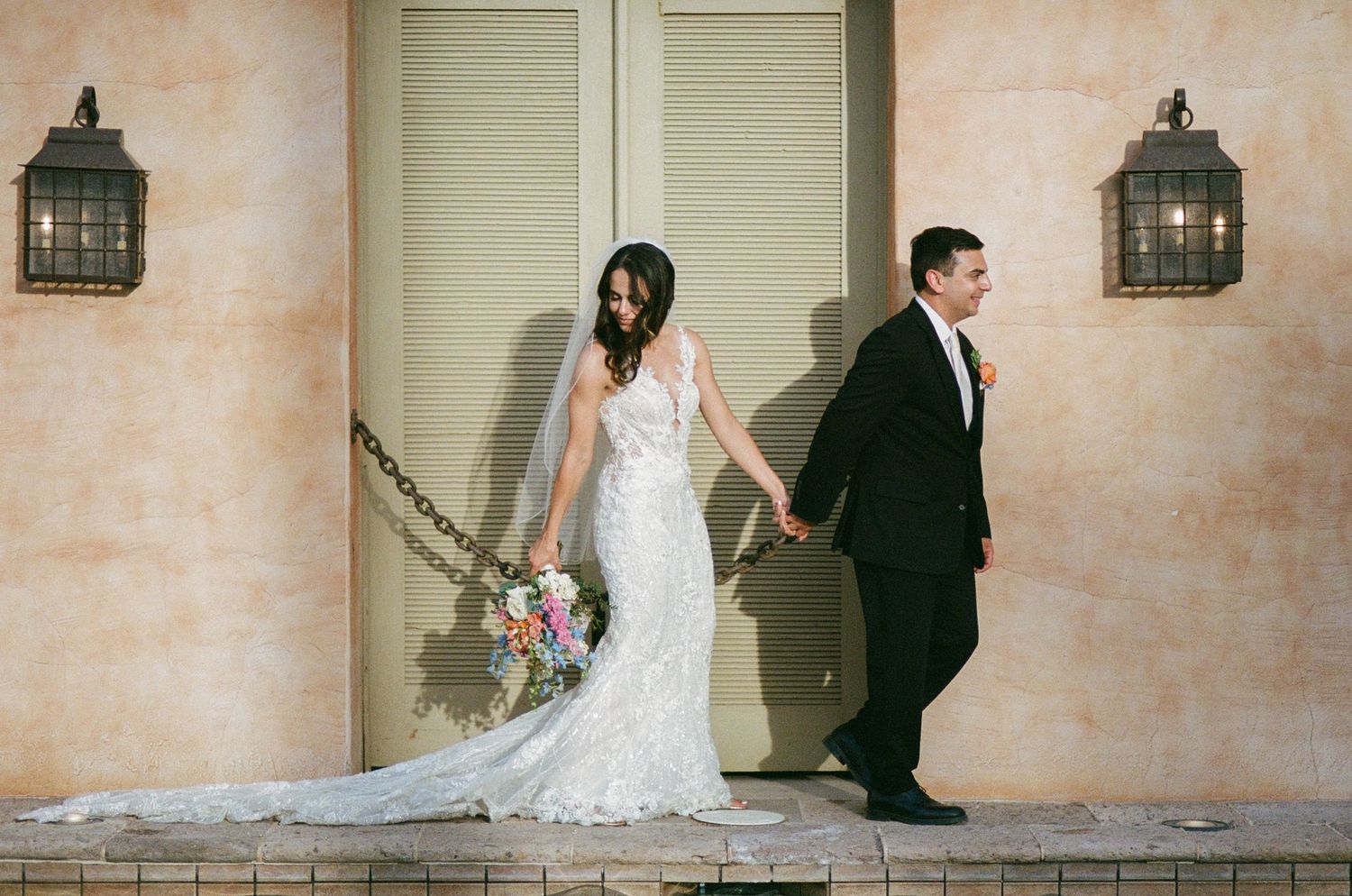 Bride in lace mermaid gown holding bouquet walks with groom in black tuxedo outside elegant venue.