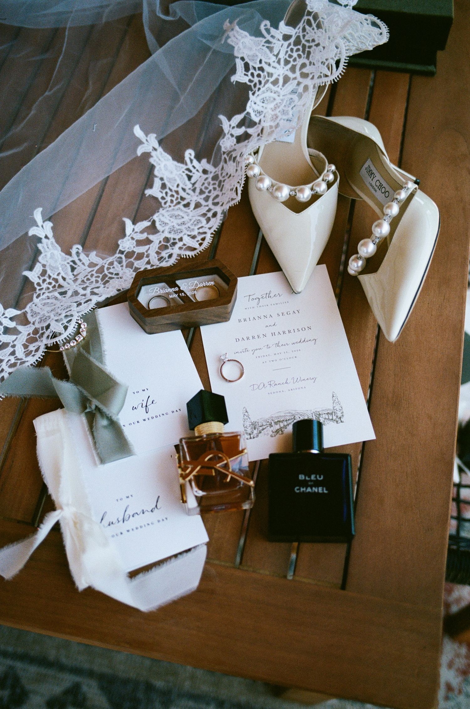 Elegant wedding flat lay with lace veil, pearl heels, Chanel perfume, invitation suite, and rings on a wooden table.