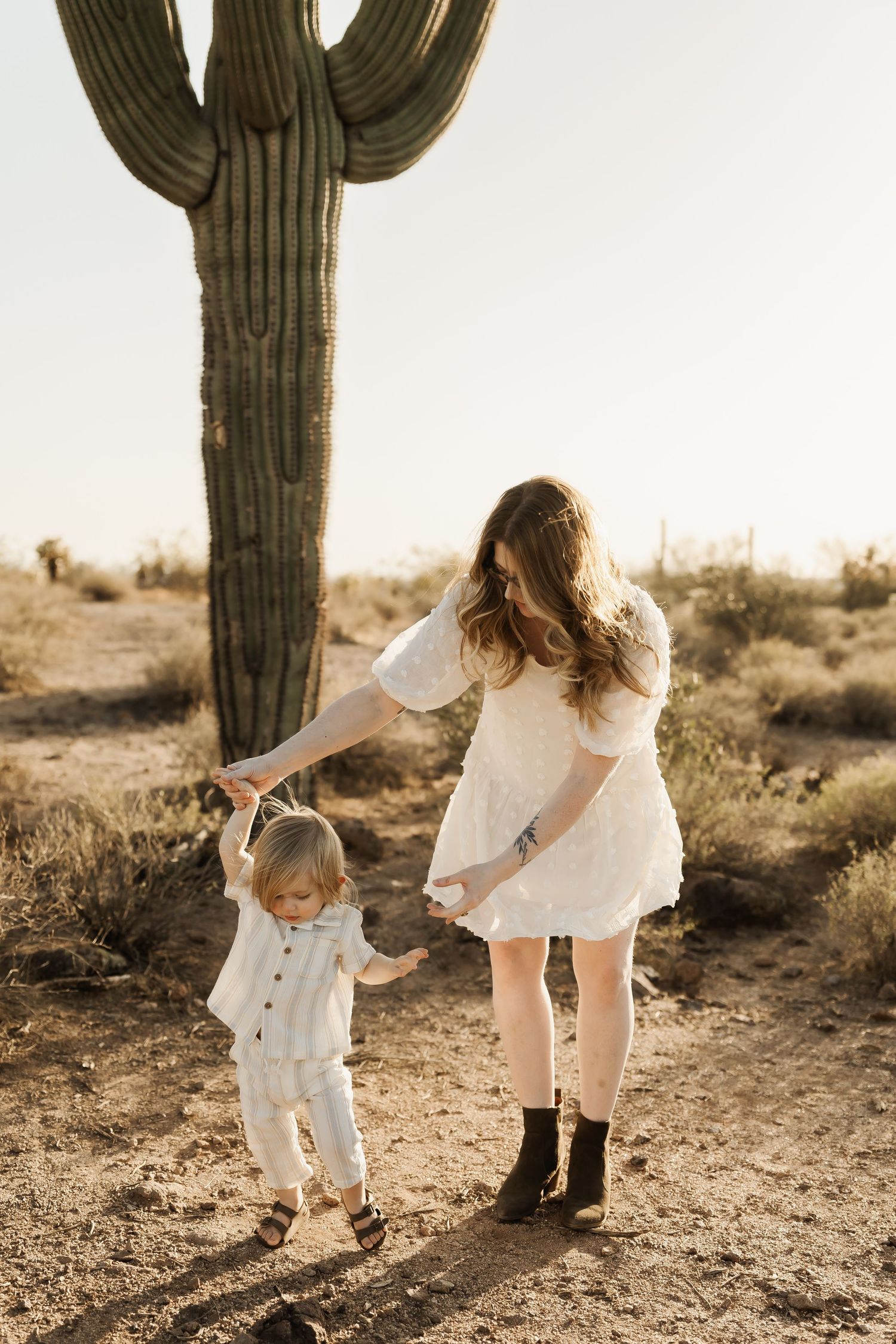 Mother and child in white outfits walking near tall saguaro cactus in desert setting.