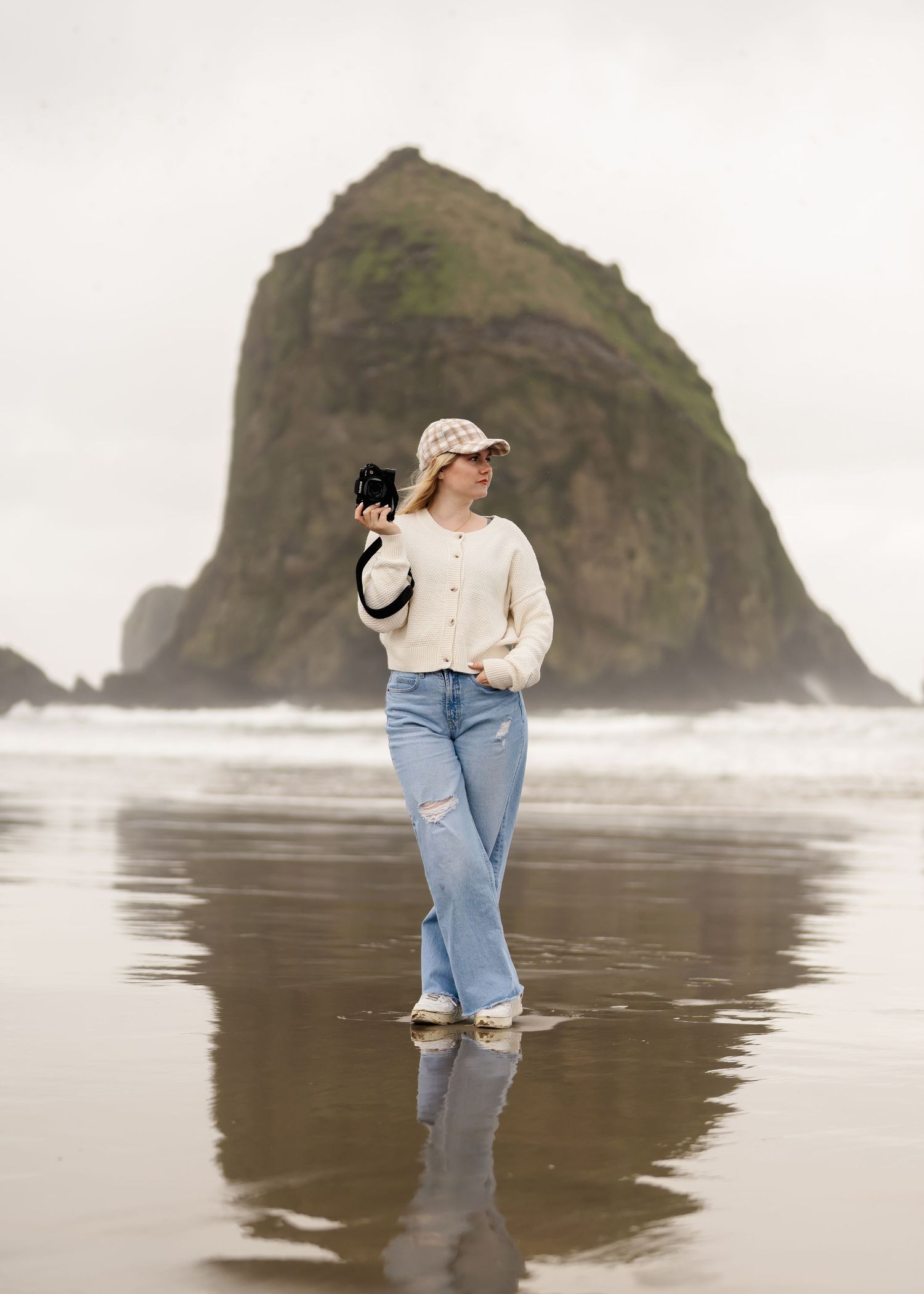 Someone in jeans and white sweater stands on reflective beach sand with Haystack Rock at Cannon Beach in background.