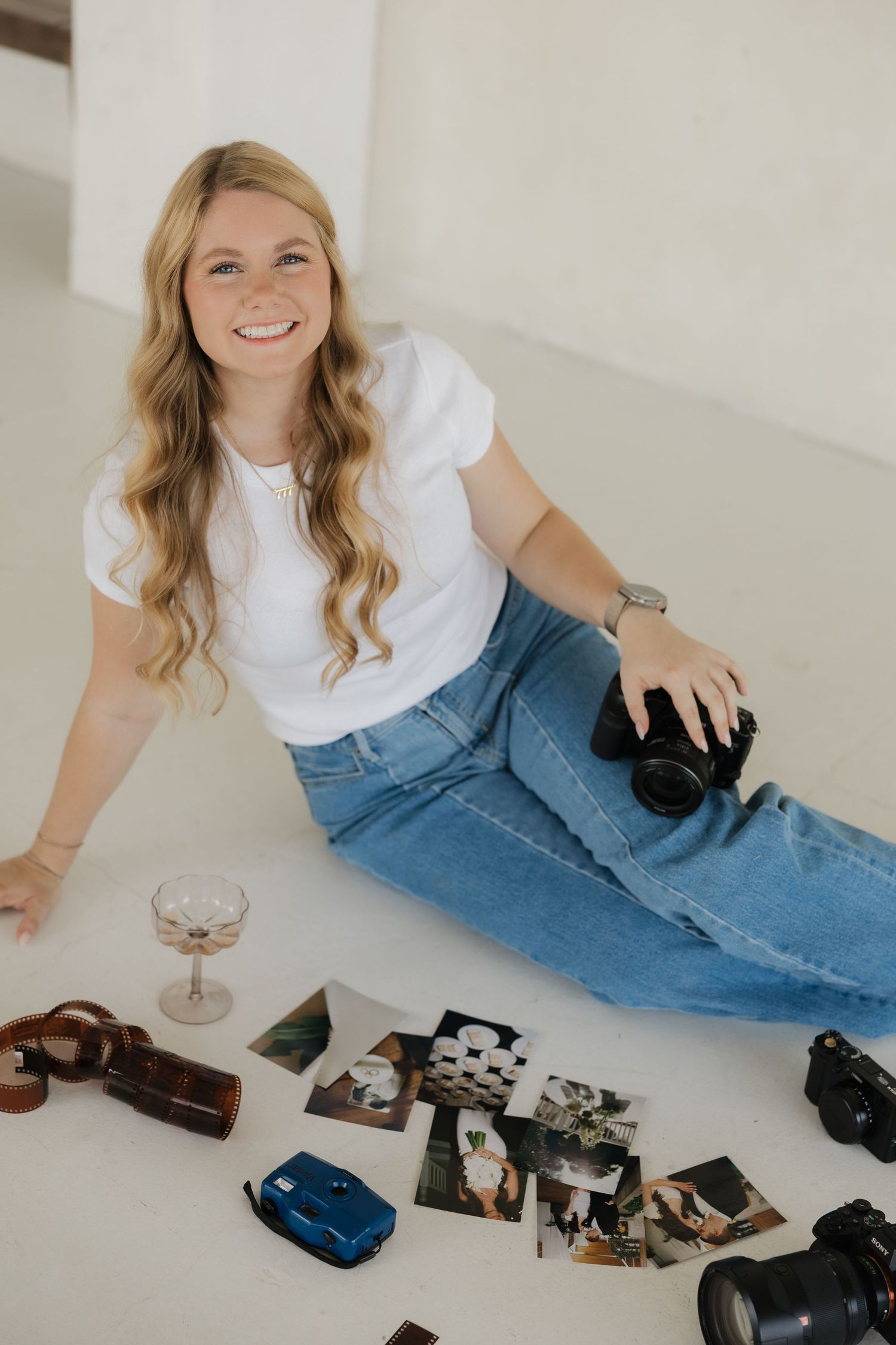 Smiling blonde woman in white tee and jeans sits on floor with cameras and photos scattered around her.