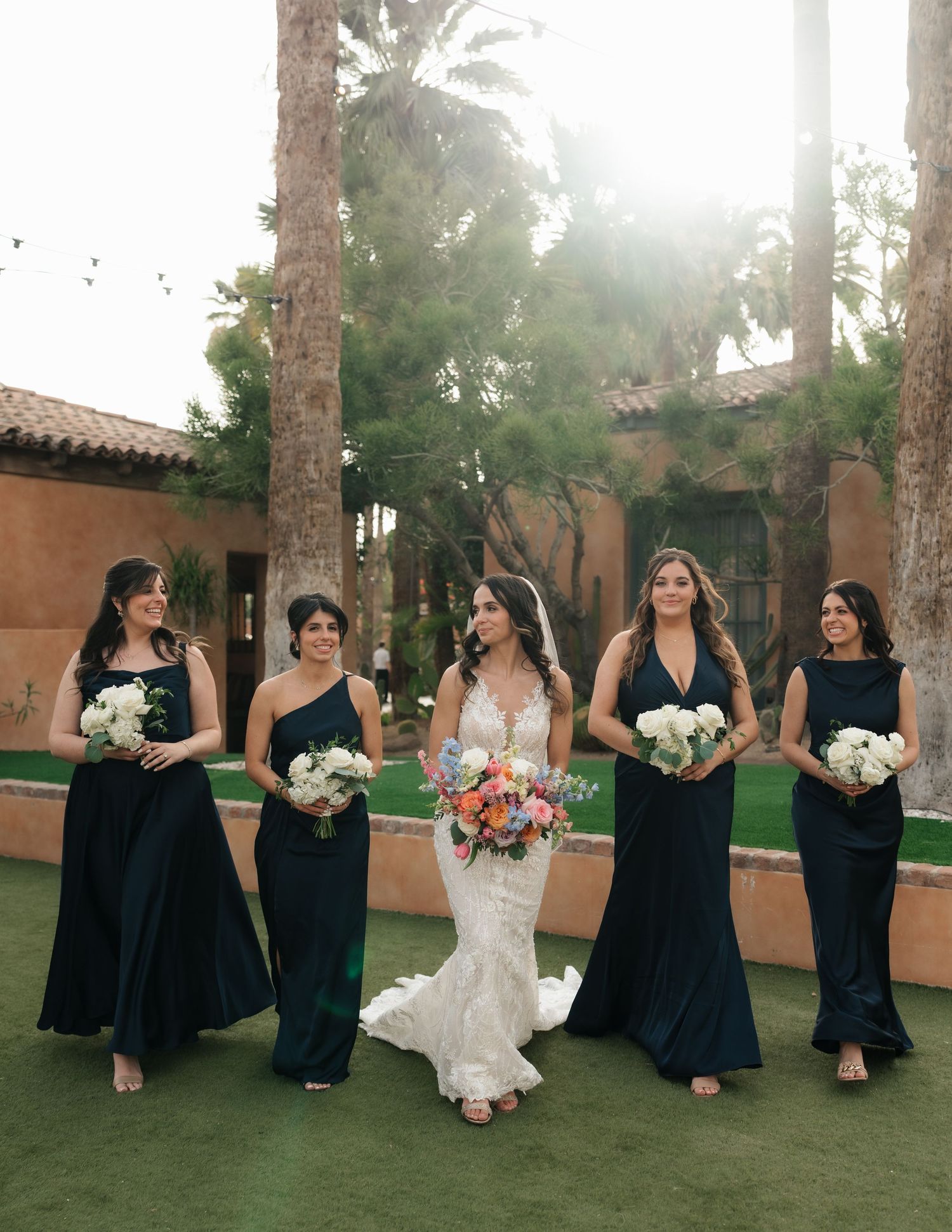 Bride in white gown walks with four bridesmaids in dark green dresses holding bouquets outdoors near palm trees.