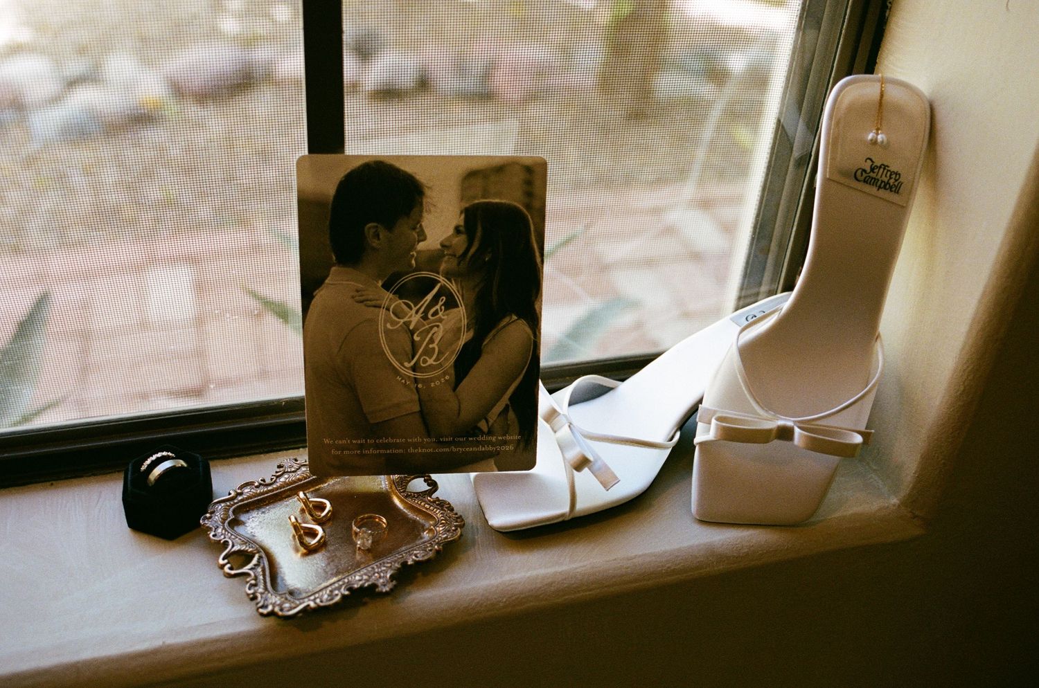 White bridal heels and engagement photo displayed on windowsill with wedding rings and gold jewelry tray.