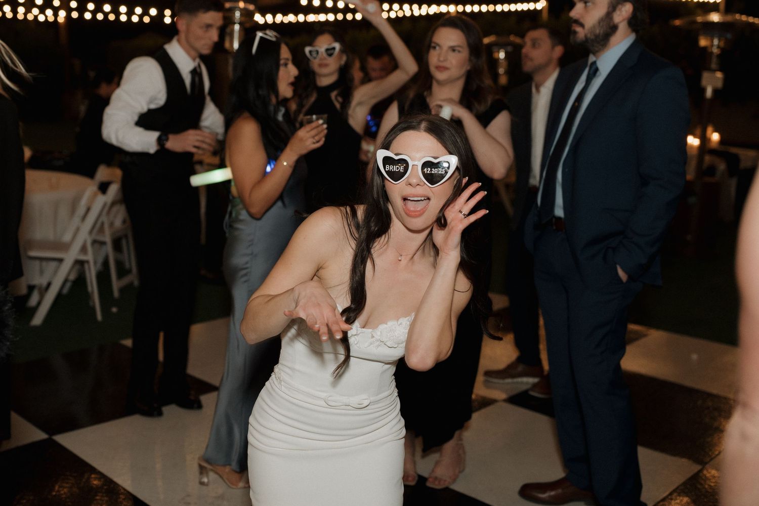 A group of people dancing and celebrating at an indoor wedding reception with string lights overhead.