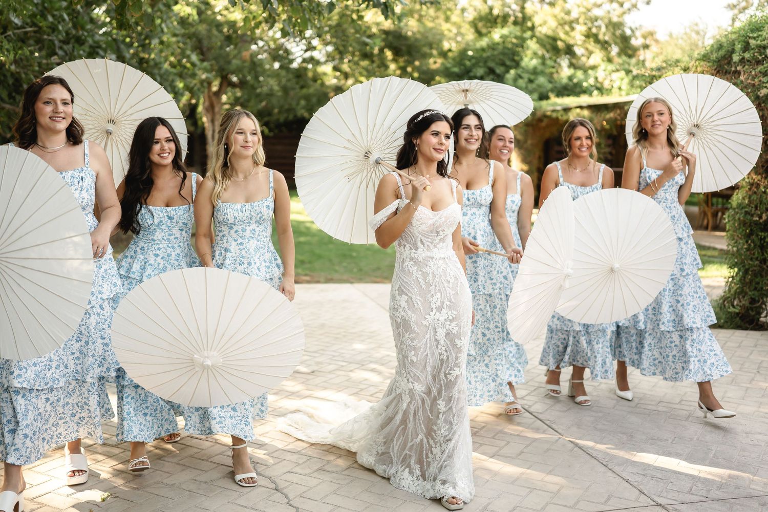 Wedding party holding white parasols while walking through a garden setting.