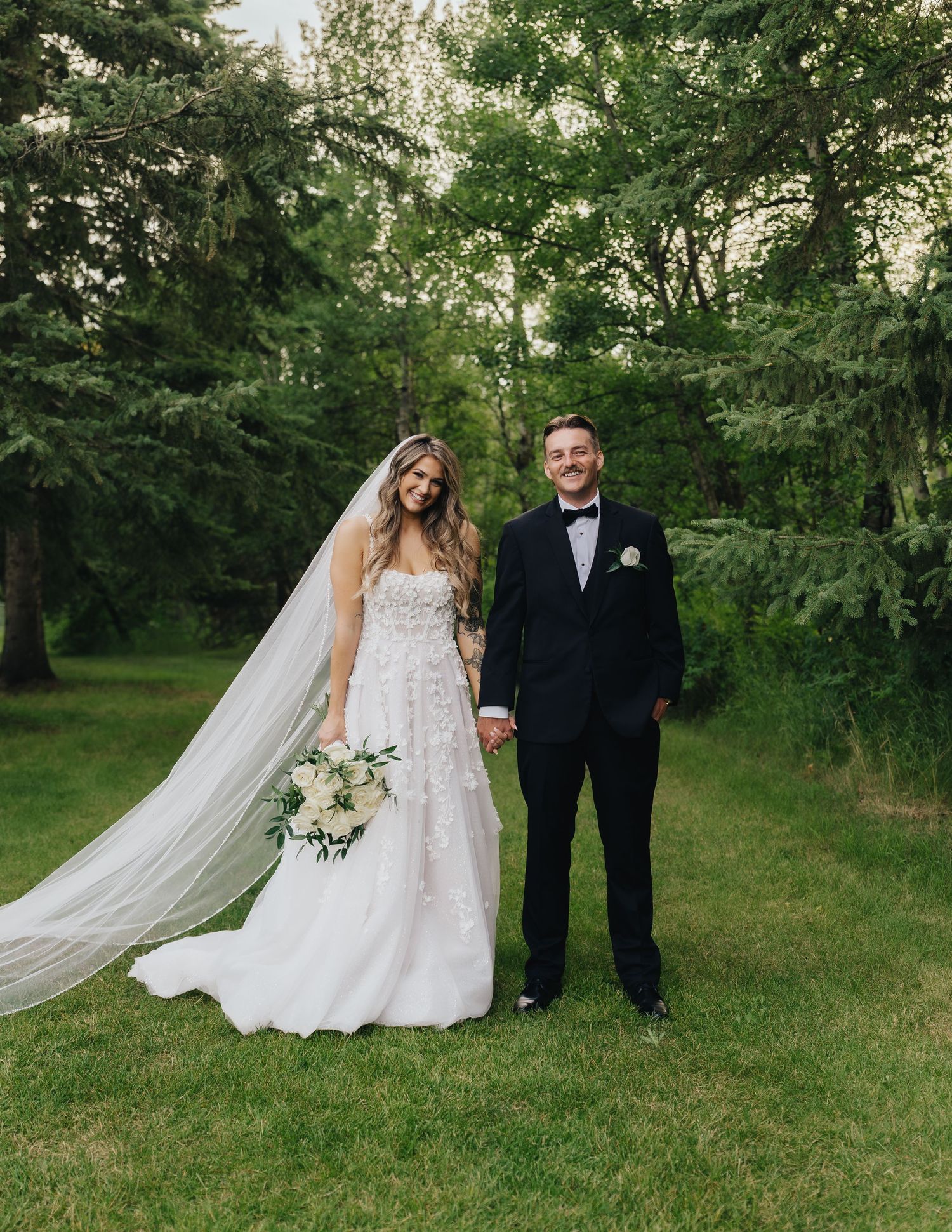 Wedding couple poses on green lawn with flowing white bridal veil in summer garden setting.