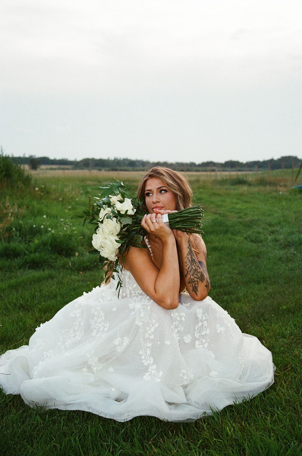 A bride in a white lace gown lays gracefully on green grass while holding a bouquet of white flowers.