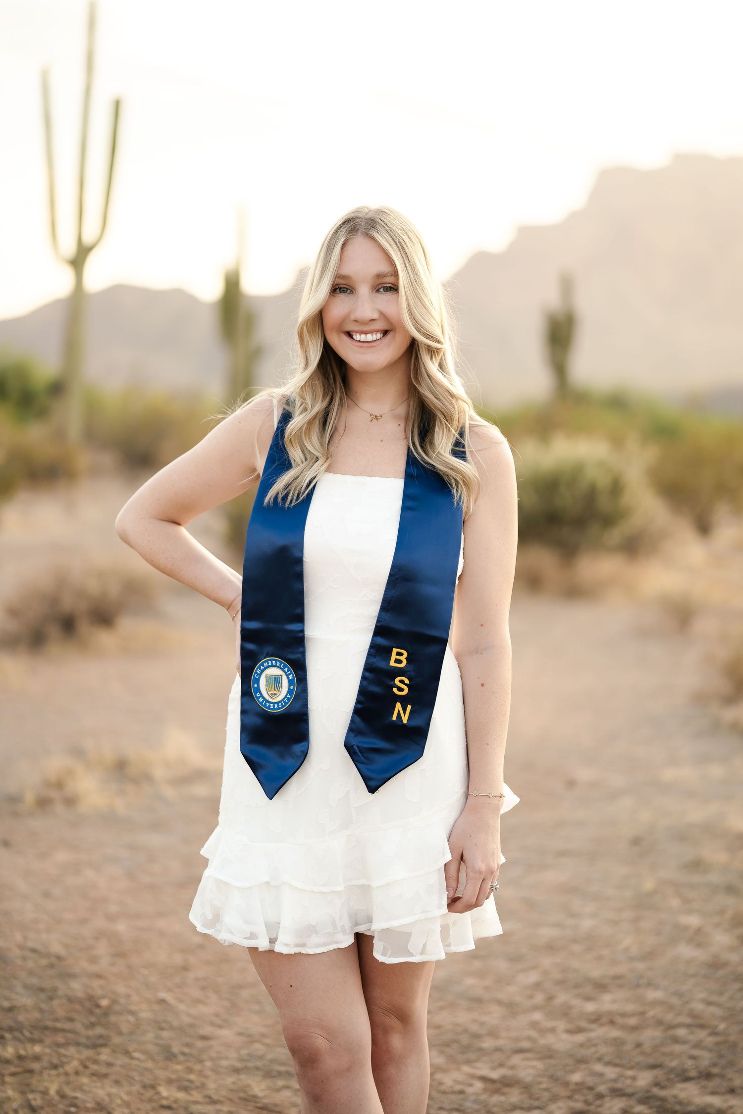 Graduate in white dress and navy stole poses confidently in desert setting with cacti.
