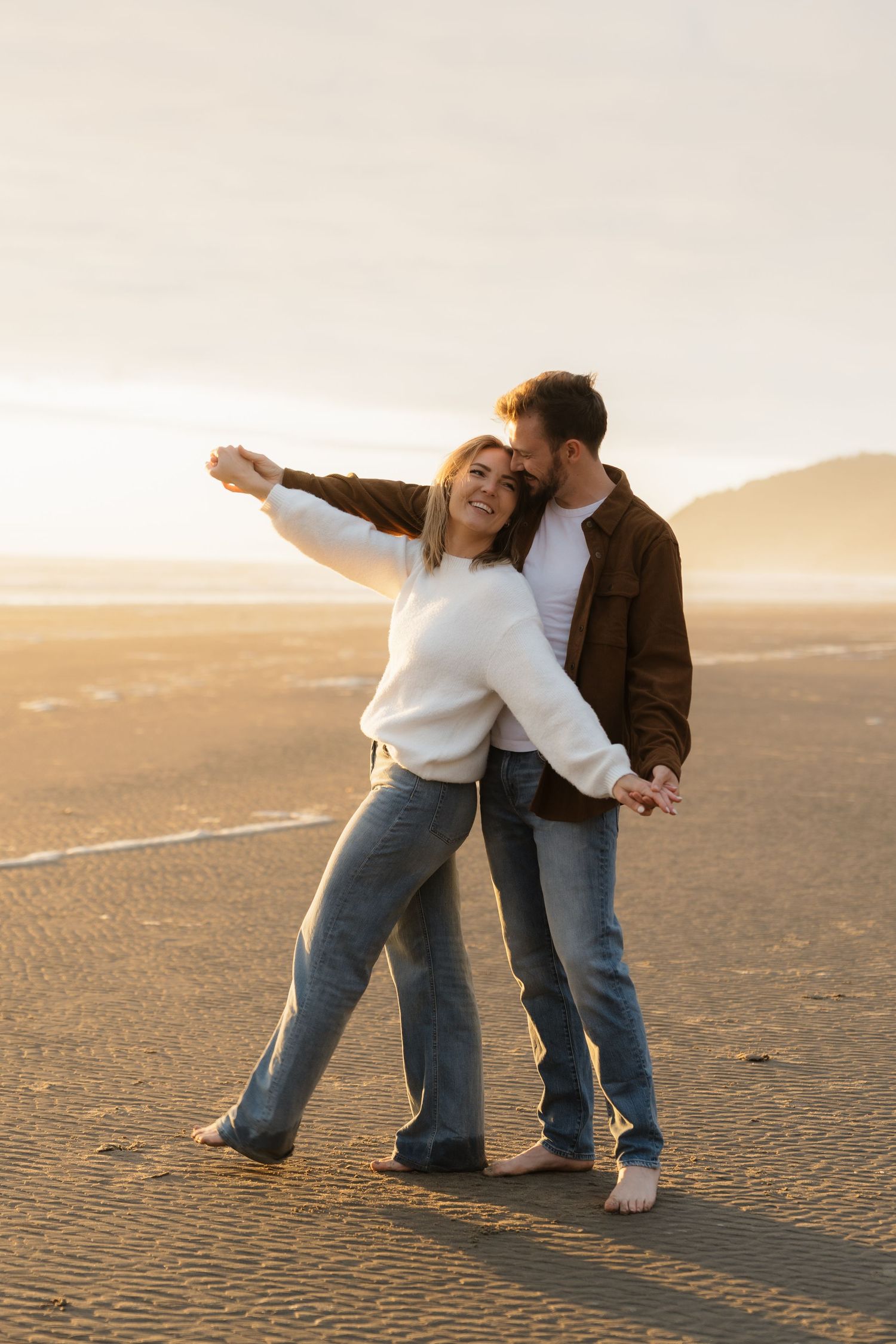 Couple dancing romantically on a sandy beach at golden sunset, woman in white sweater and jeans, man in brown jacket.