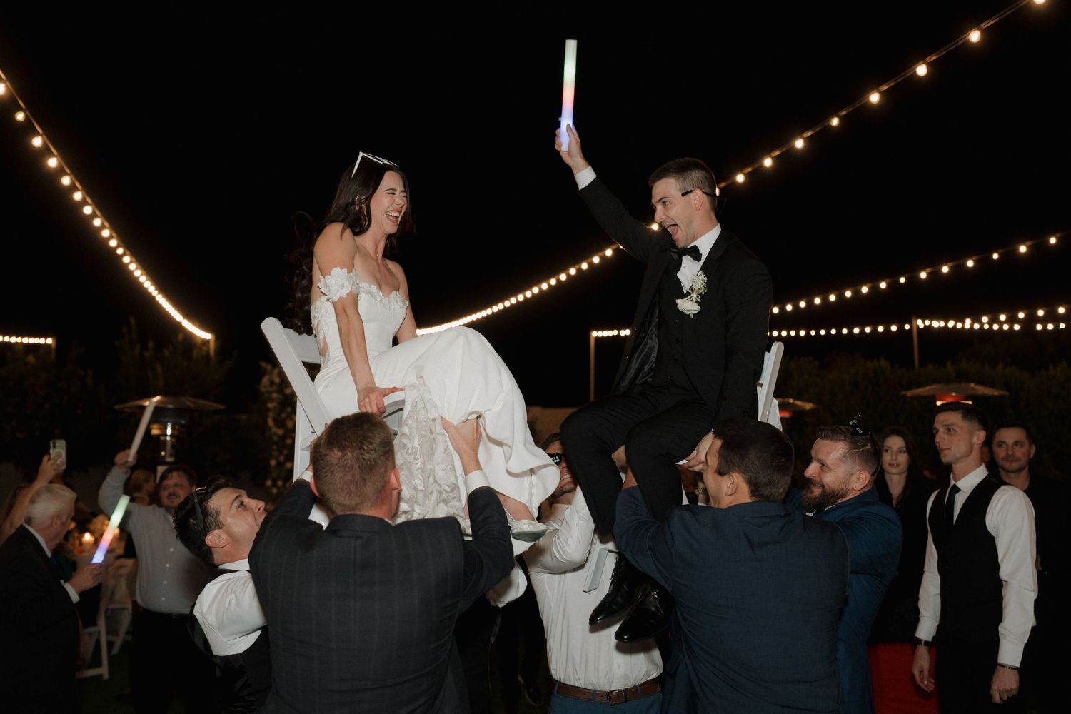 A wedding celebration at night with a couple being lifted on chairs during the hora dance under string lights.