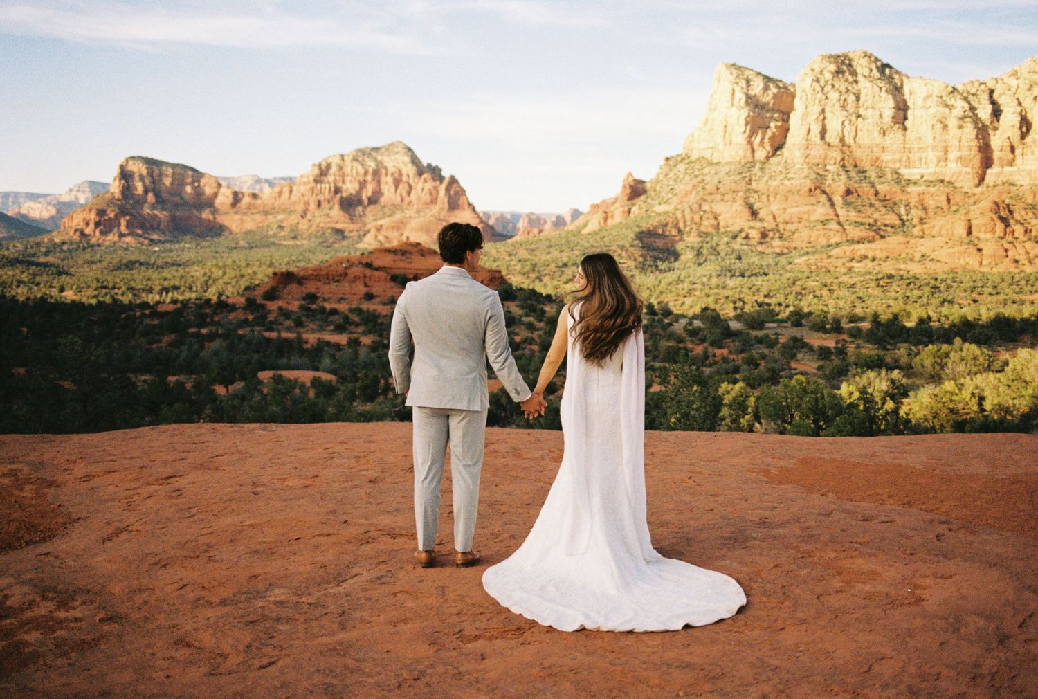 Couple in wedding attire holding hands overlooking stunning Sedona red rock landscape at golden hour.