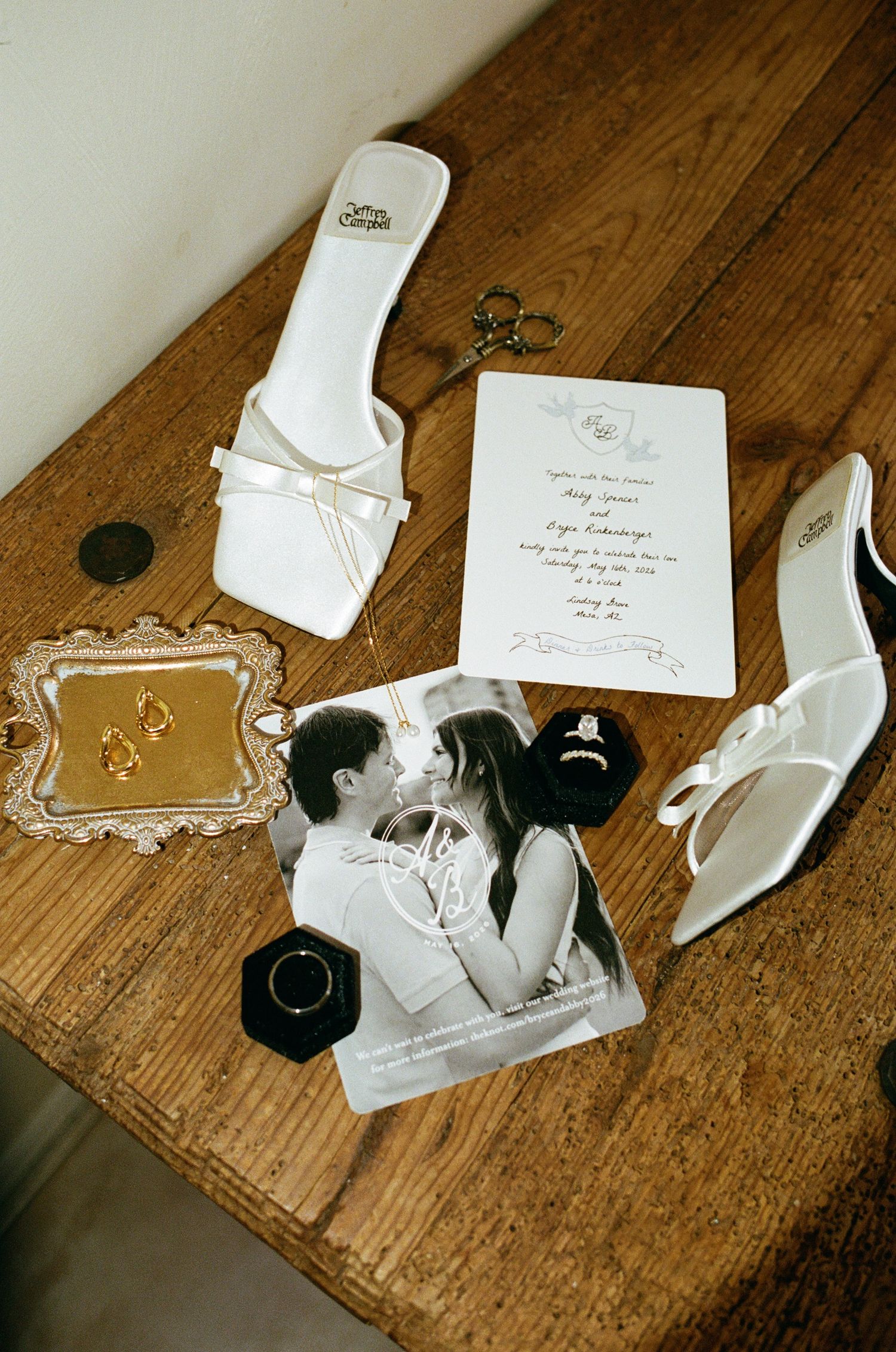 White bridal heels, wedding invitation, engagement ring, gold earrings, and couple photo styled on rustic wood table.