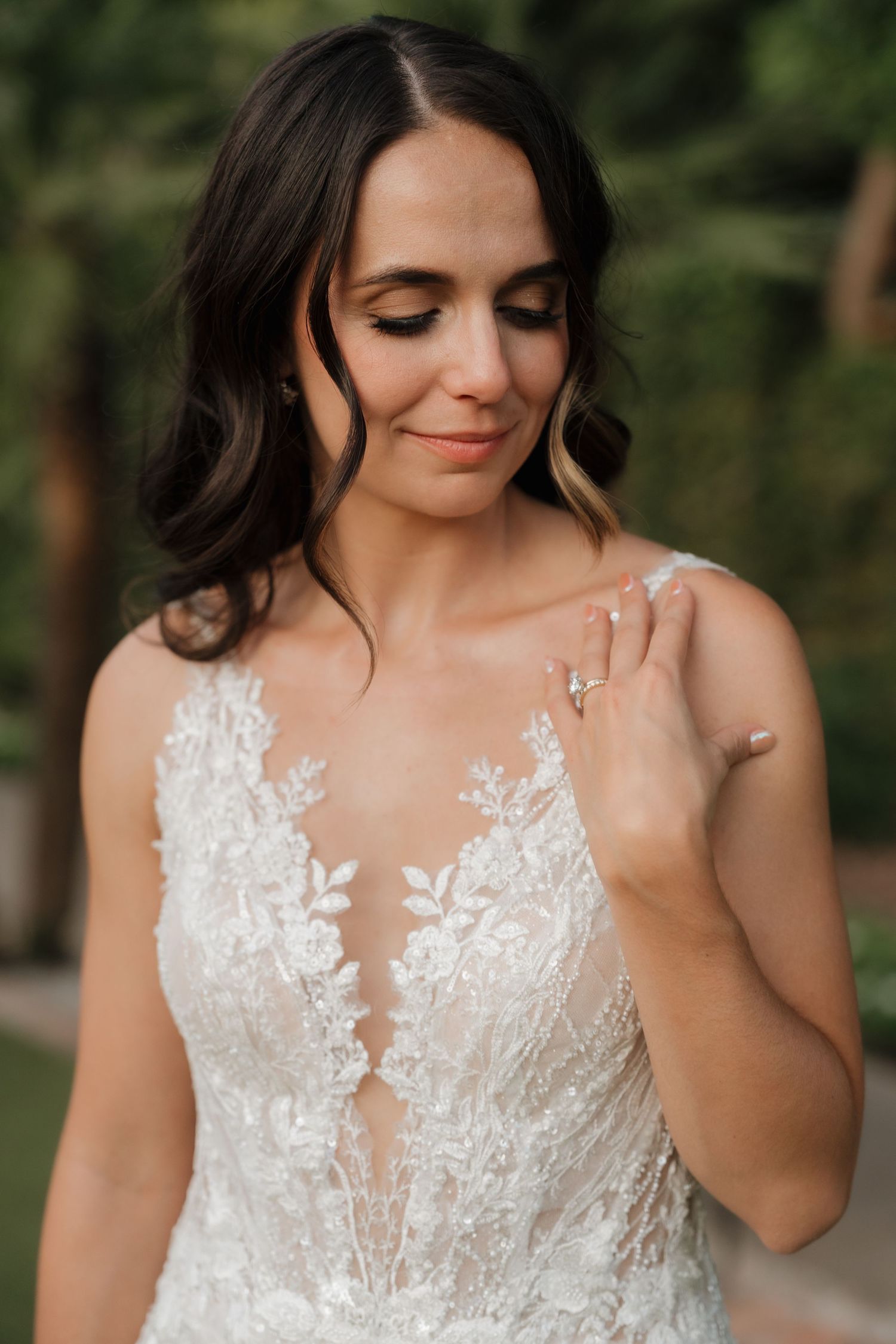 Bride in lace illusion wedding gown smiling downward, showcasing elegant diamond engagement ring outdoors.