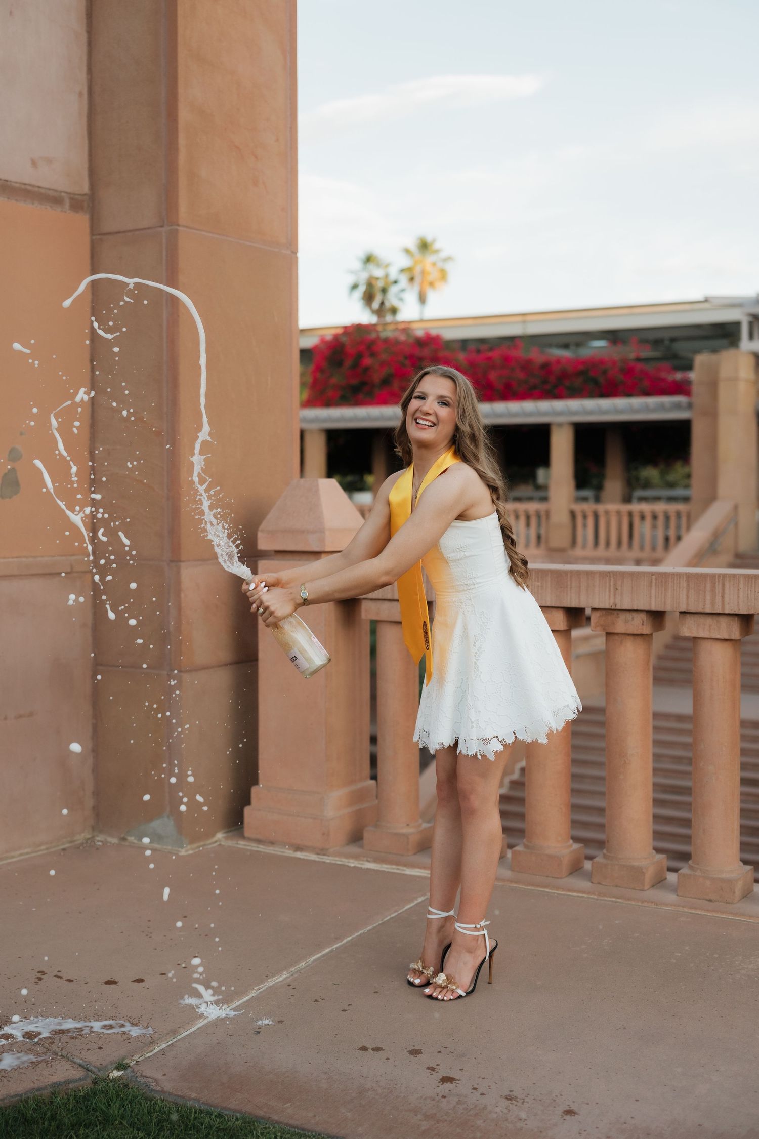 Young woman in white dress and yellow sash joyfully popping champagne bottle outdoors at elegant venue.