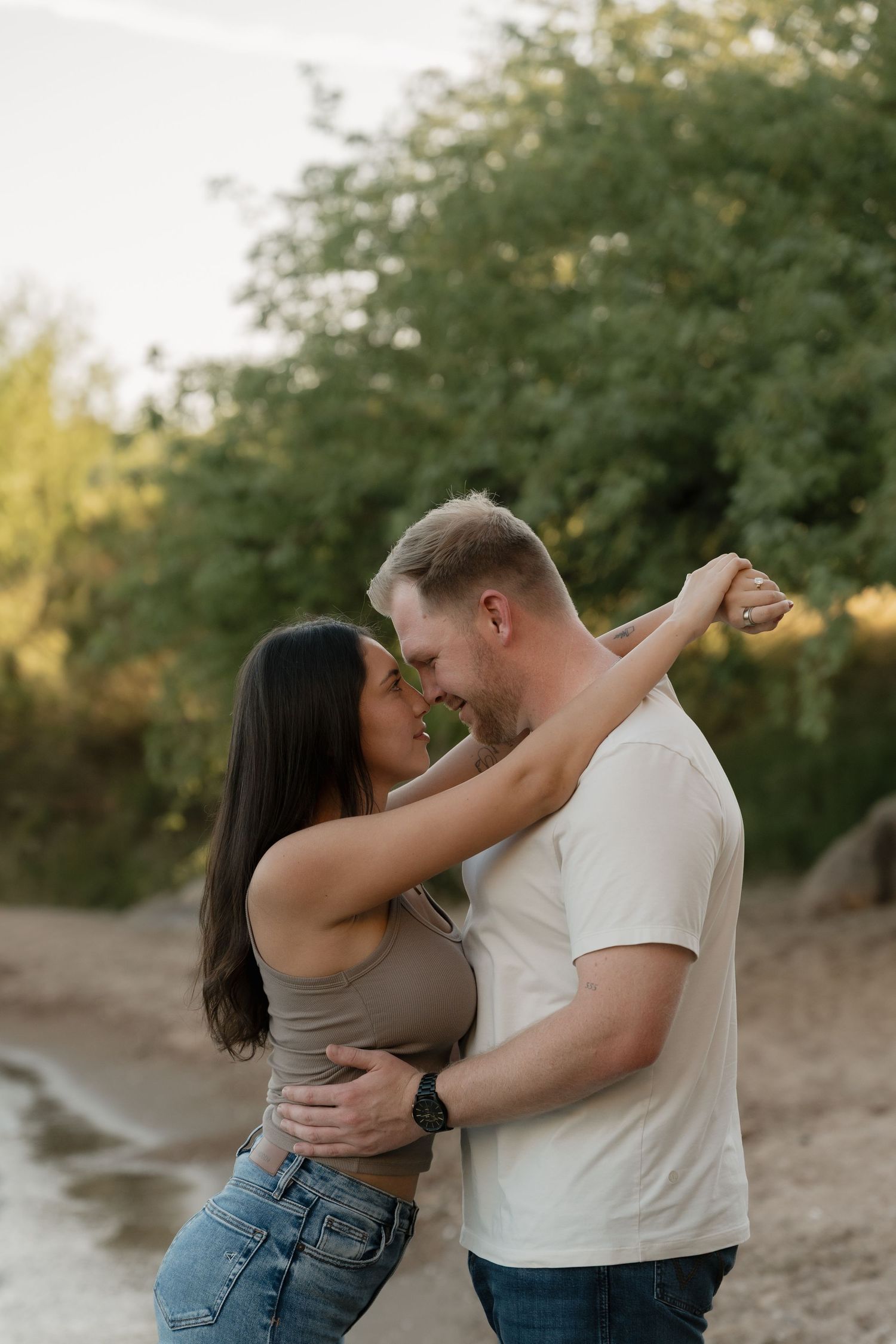 A couple embraces and leans in for a kiss outdoors, surrounded by lush green trees on a sandy path.
