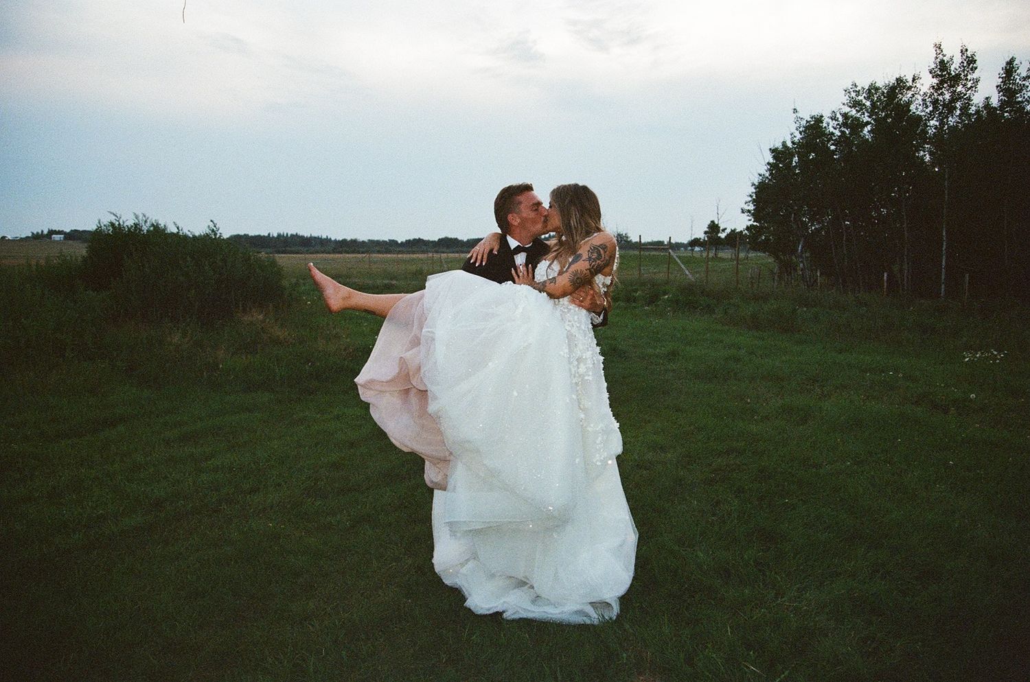 Groom lifting bride in wedding dress for a kiss outdoors in a lush green field on their wedding day.