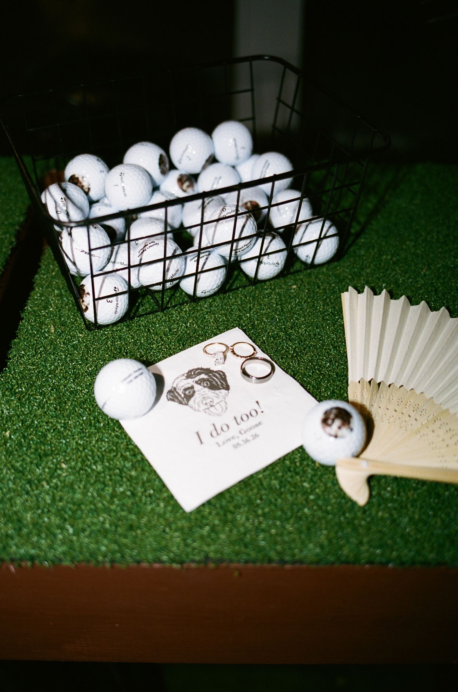 Custom golf balls in a wire basket with wedding program, rings, and a fan on artificial turf at a golf-themed wedding.