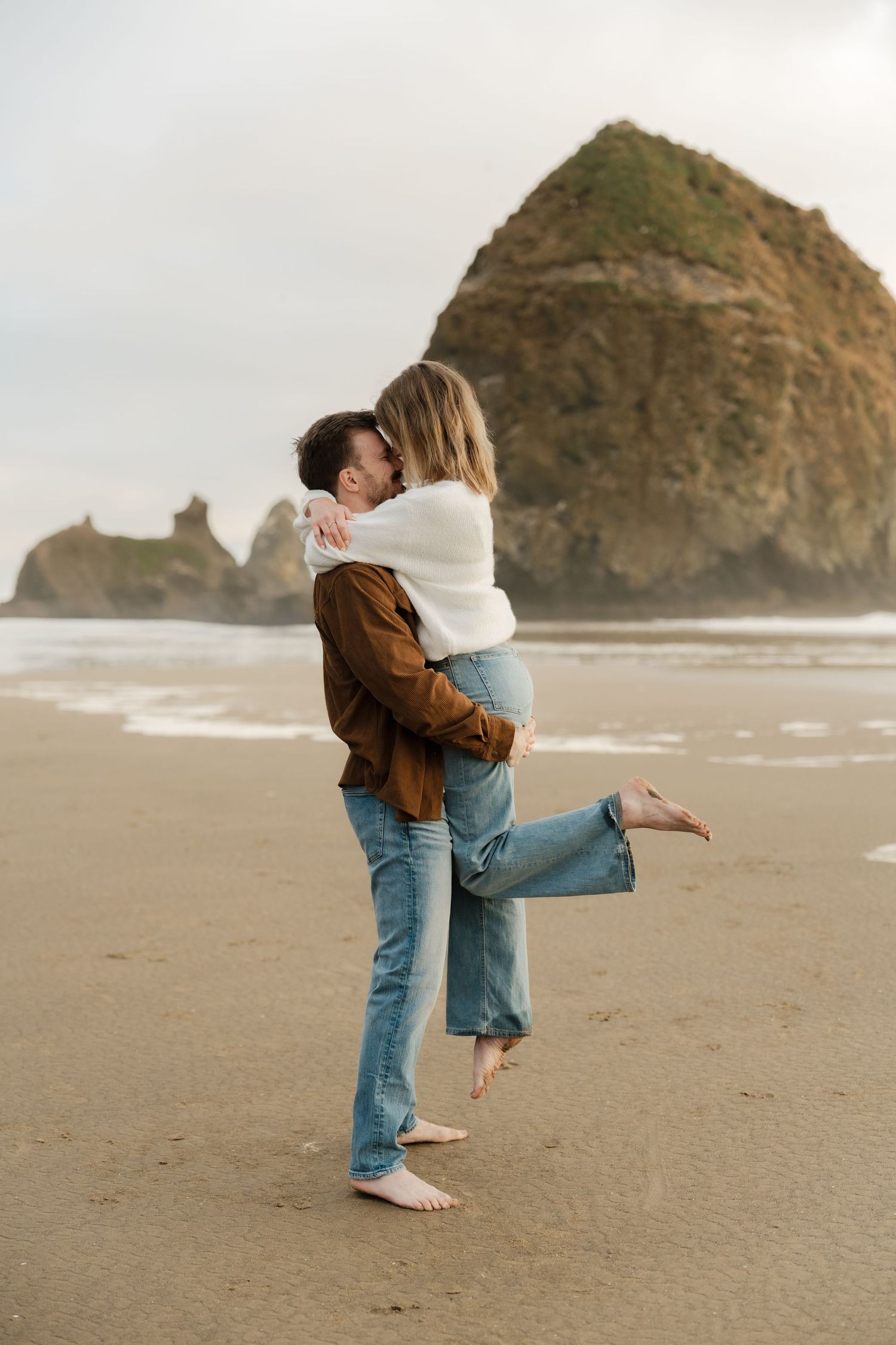 Couple embracing on a sandy beach with Haystack Rock in the background on an overcast day.
