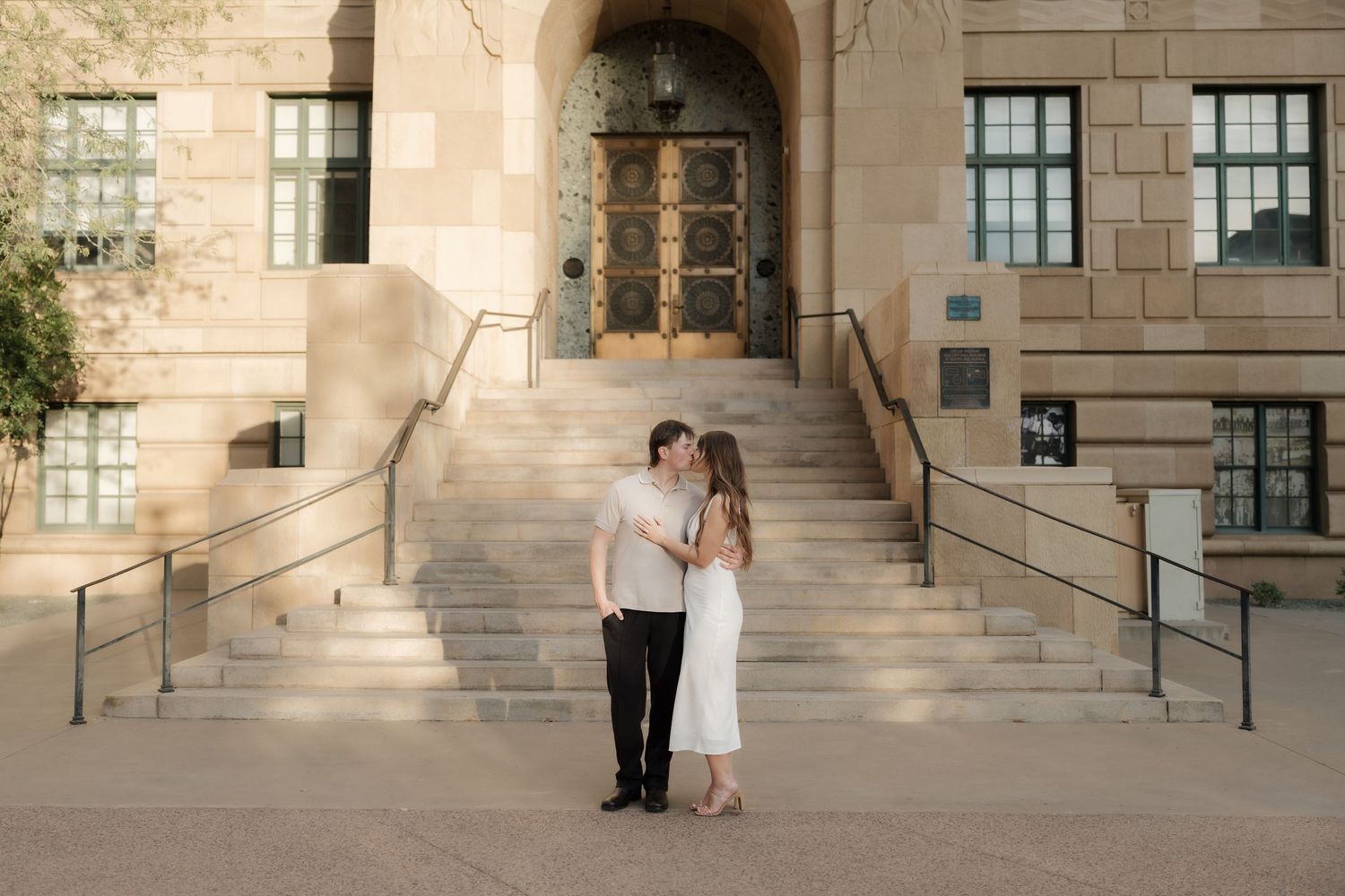 A couple standing on the steps of a stone building, with arched doorways and windows in the background.