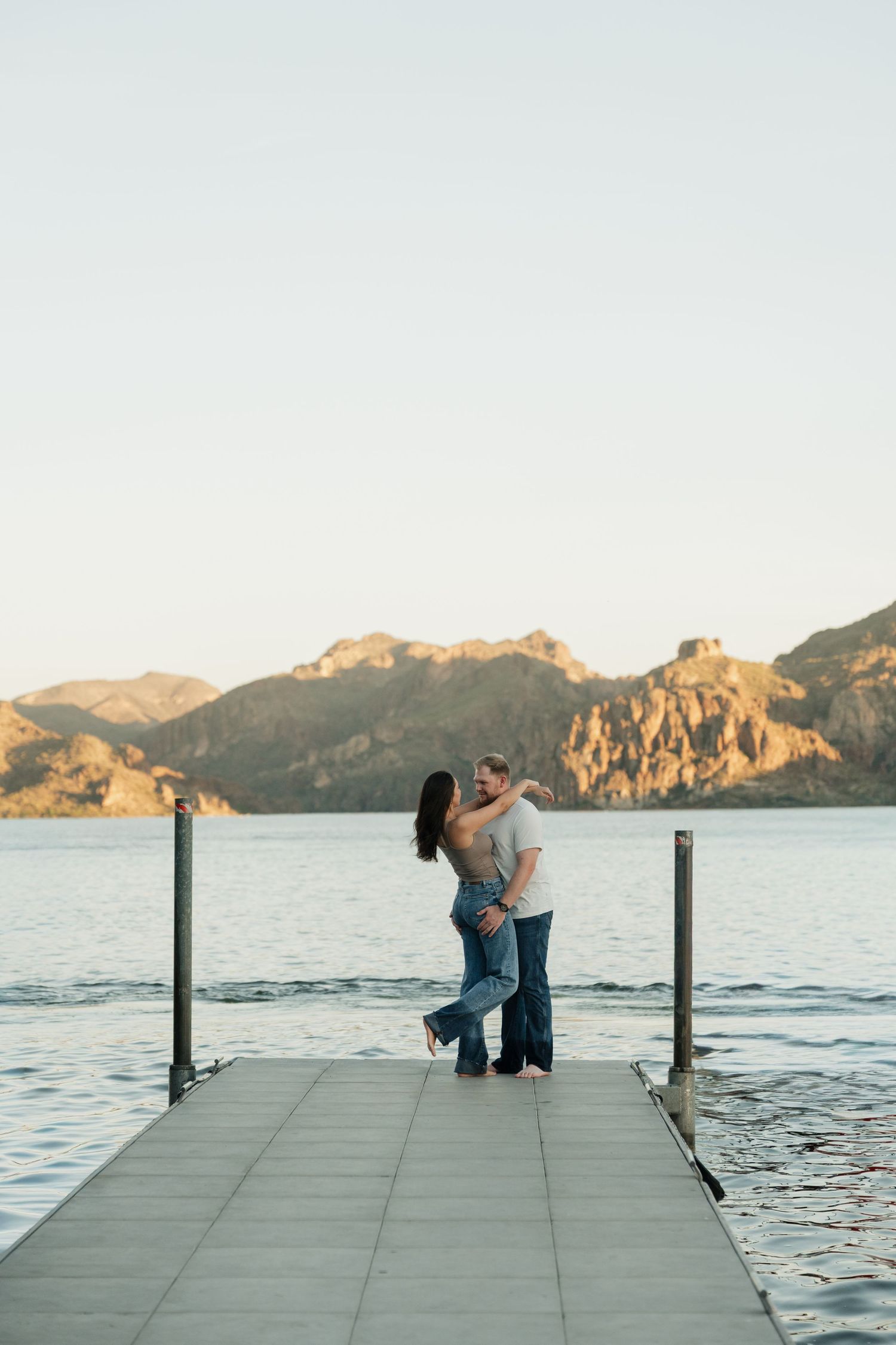 Couple embracing on a dock at sunset with mountains and calm lake in the background.