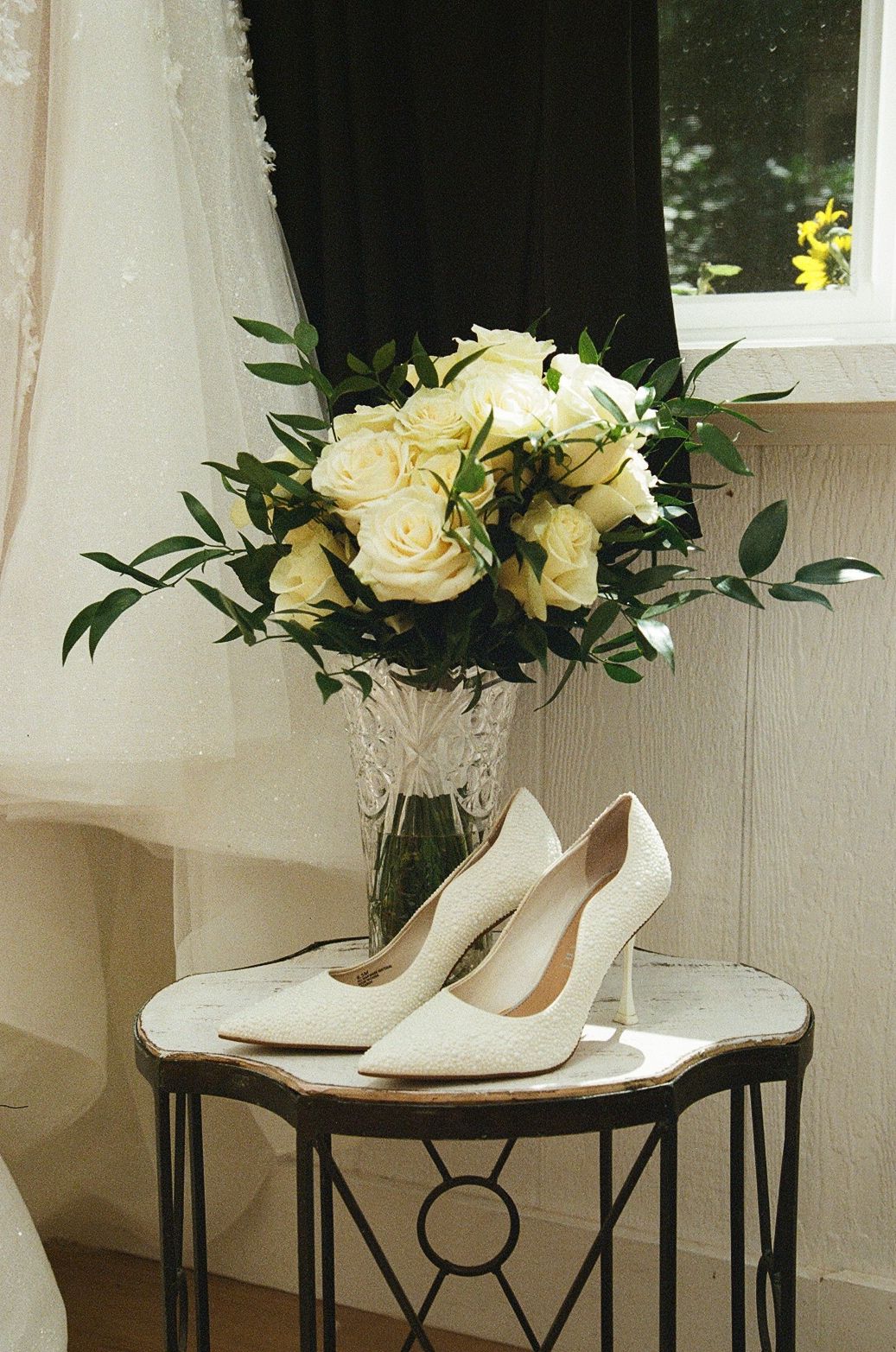 White bridal heels and white rose bouquet in crystal vase on ornate side table near window.