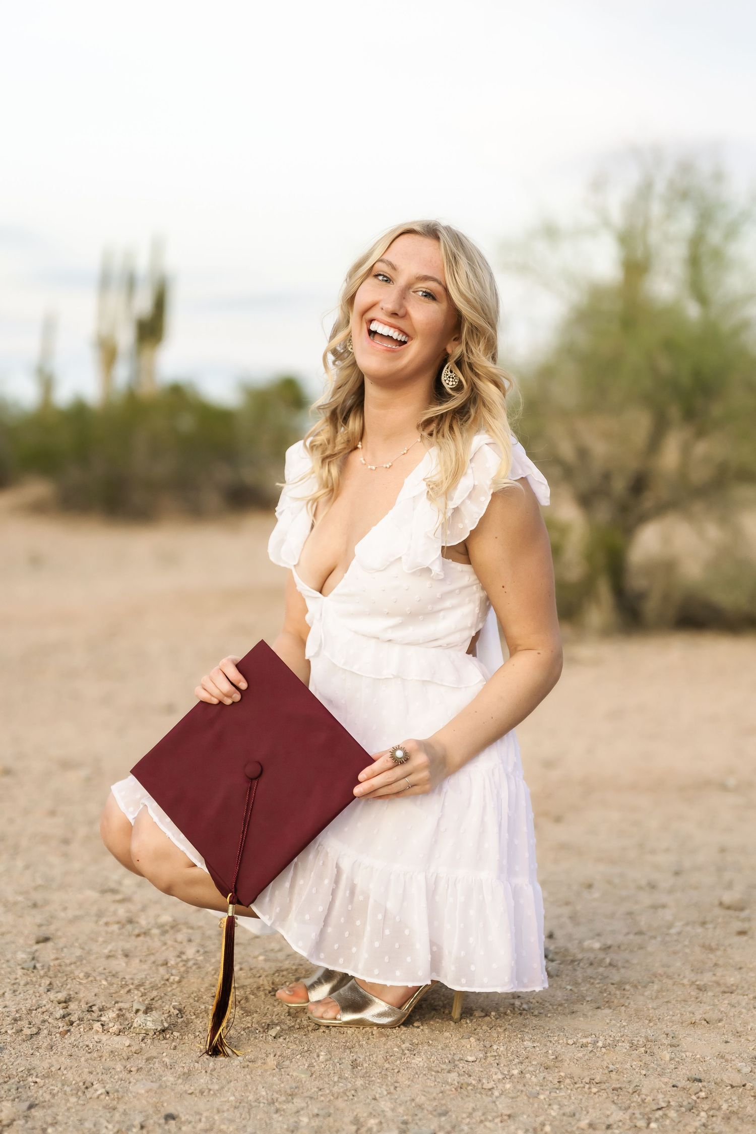 Graduate in white dress poses with diploma on desert ground.