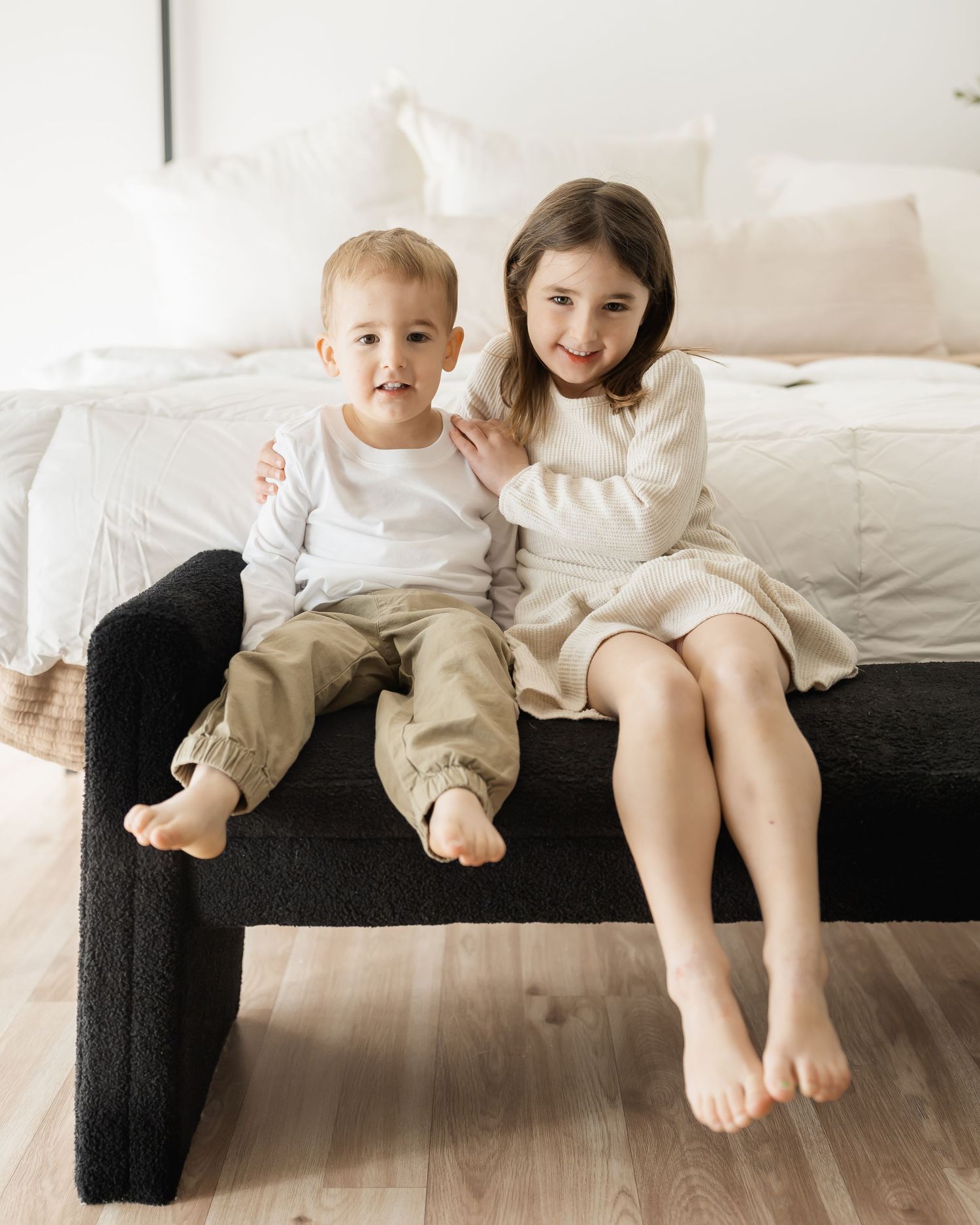 Young siblings showing their bare feet while sitting together on a dark couch in a bright room.
