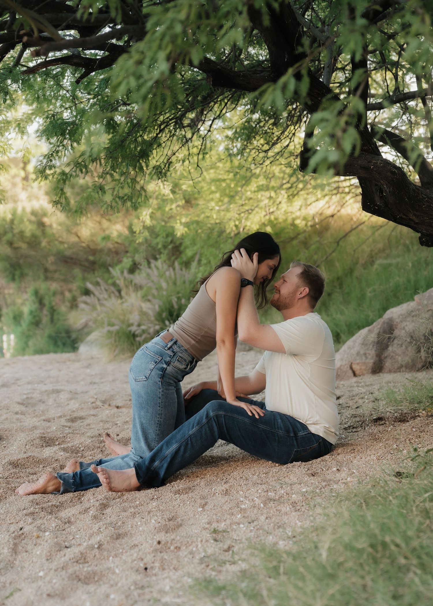 Couple sharing a romantic moment sitting on sandy ground under a lush green tree outdoors.