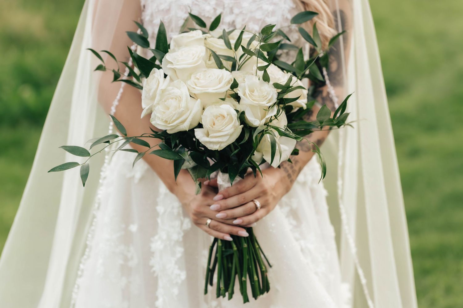 A wedding bouquet of white roses and greenery held against a white lace dress in an outdoor garden setting.