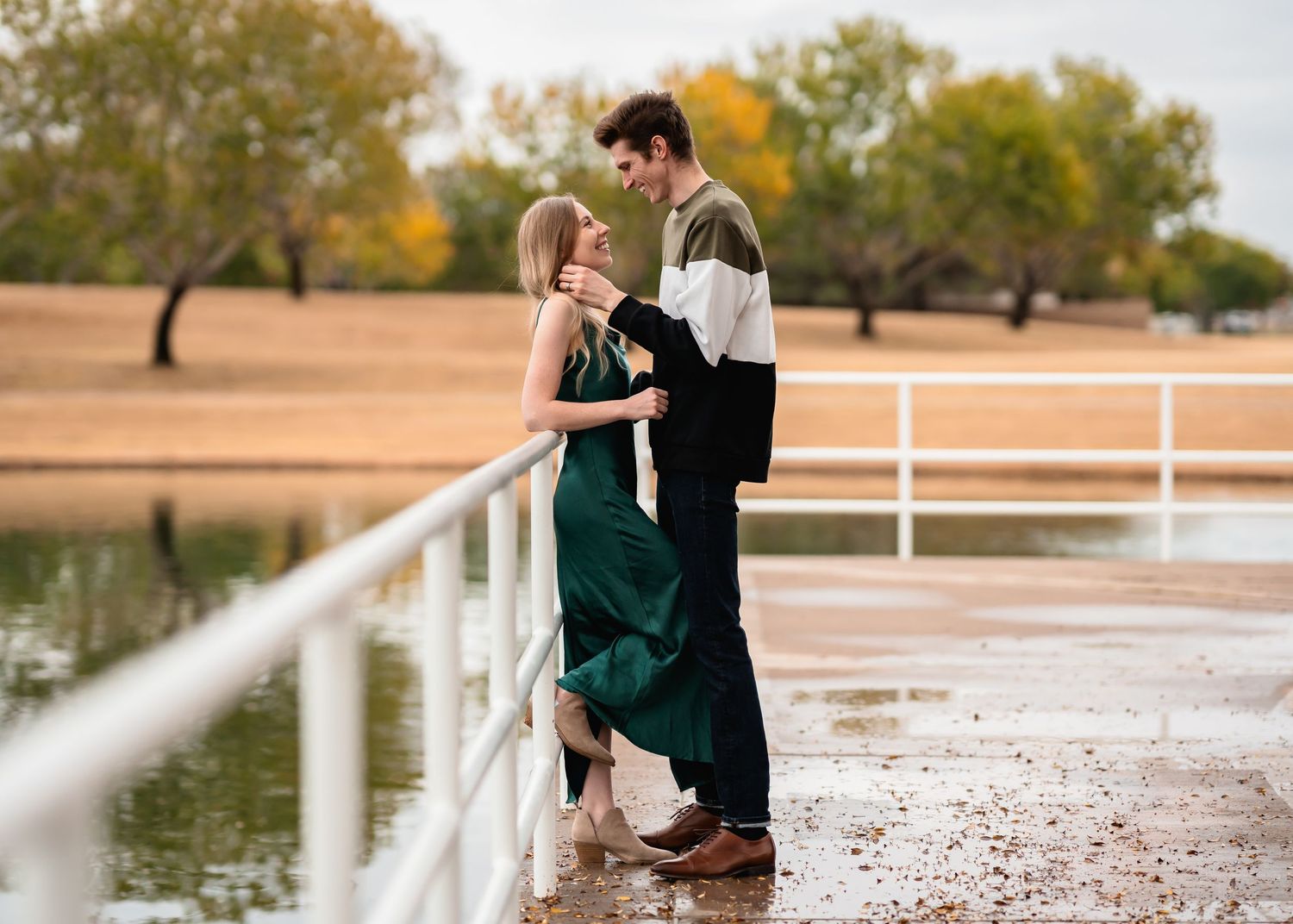 Romantic couple embraces in fall scenery by a lake with white railings during sunset.