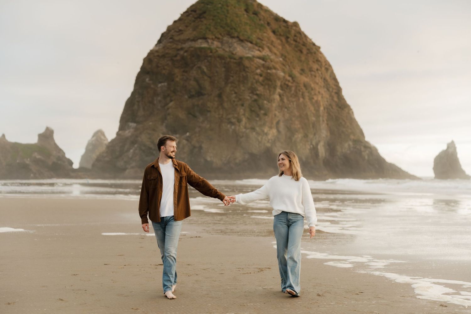 Couple holding hands walking on a beach with Haystack Rock in the background at Cannon Beach, Oregon.