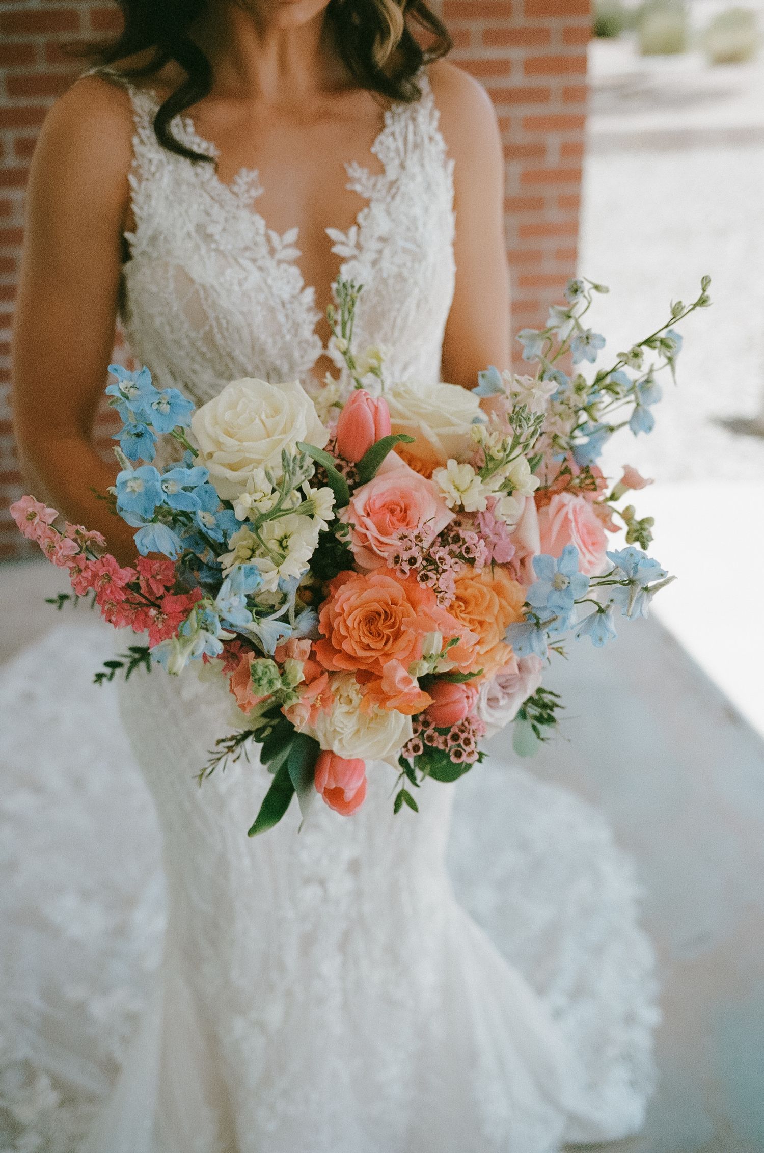 Bride in lace wedding gown holding a lush bridal bouquet with peach roses, blue delphinium, and pink blooms.
