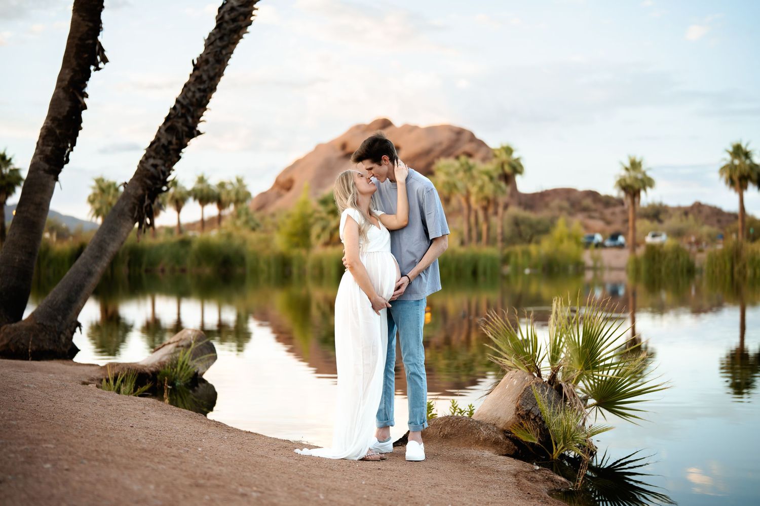 A romantic couple embraces by a serene lake at sunset surrounded by palm trees and desert landscape.