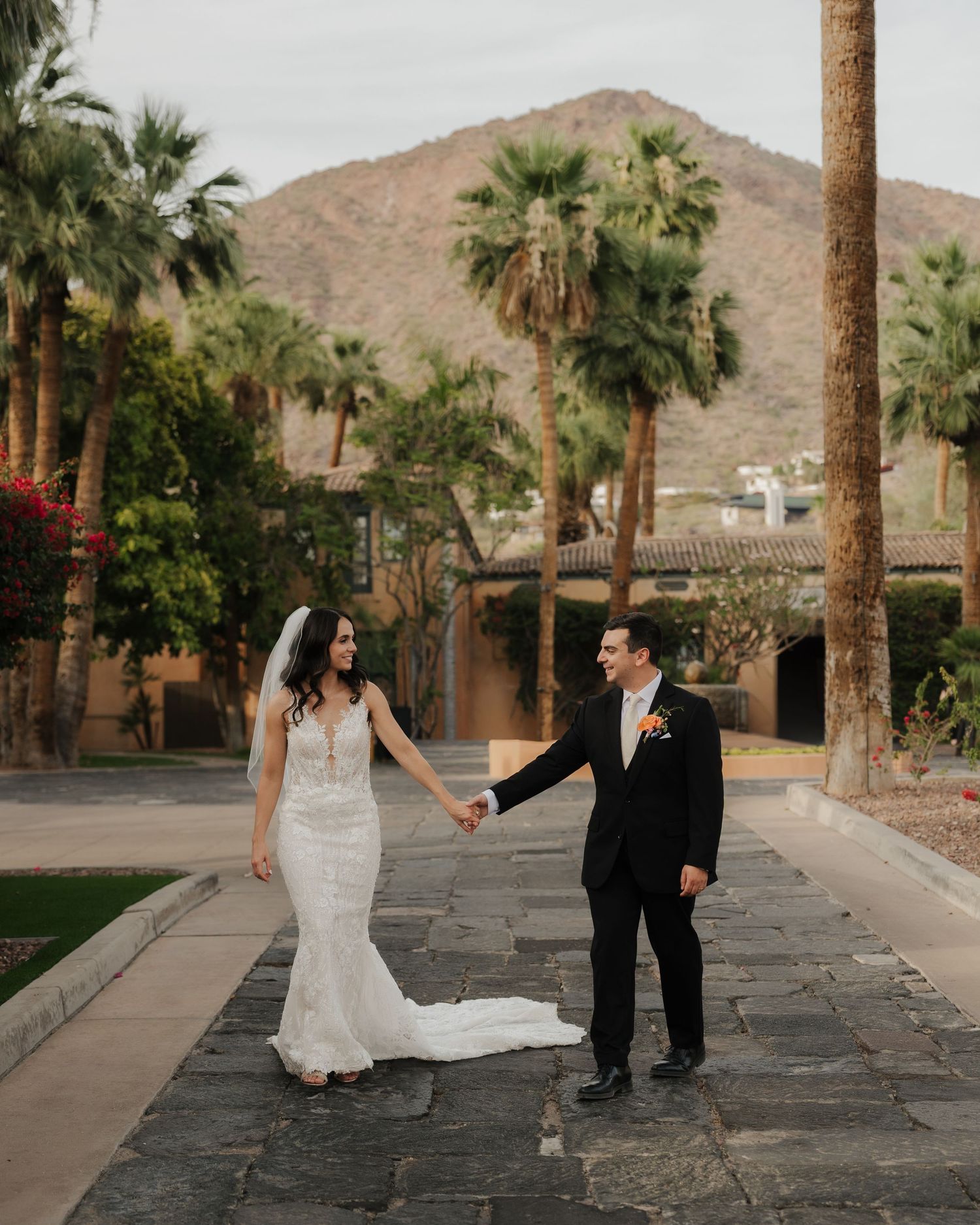 Bride and groom hold hands on stone path with palm trees and mountain backdrop at outdoor wedding venue.