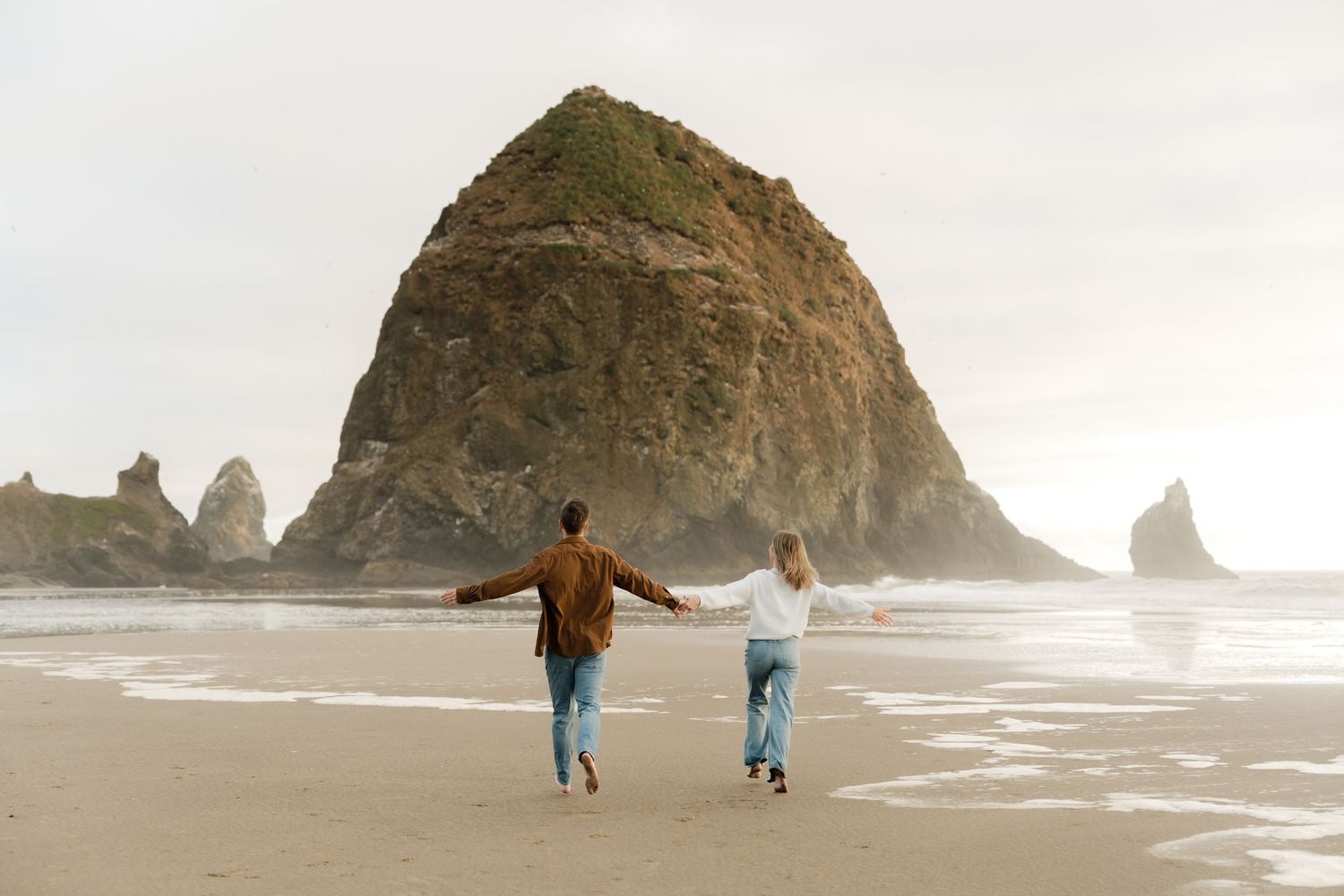 Couple holding hands running on Cannon Beach with iconic Haystack Rock in the background on a cloudy day.