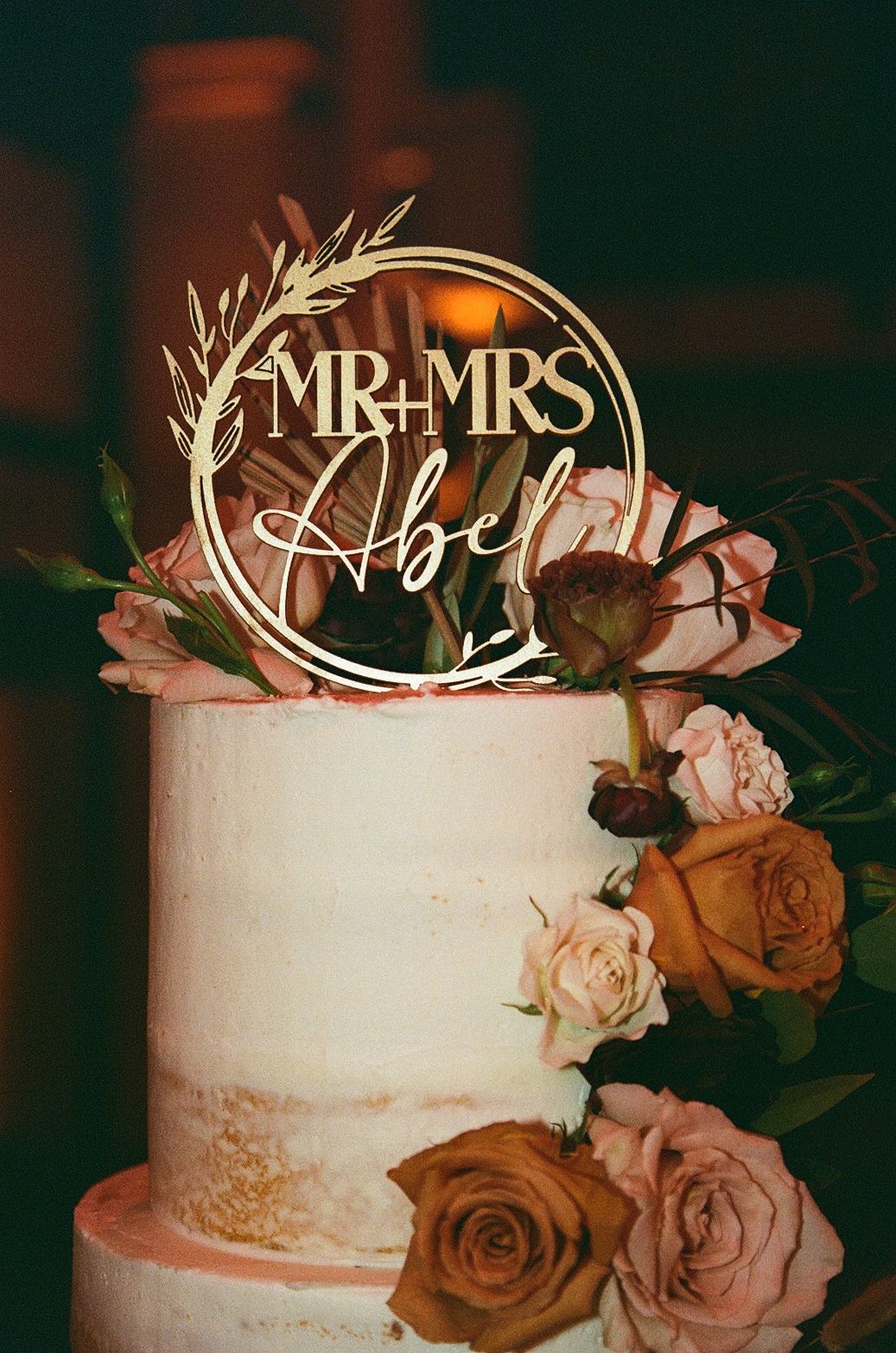 Gold Mr & Mrs Abel wedding cake topper on white tiered cake decorated with pink, orange, and burgundy roses.