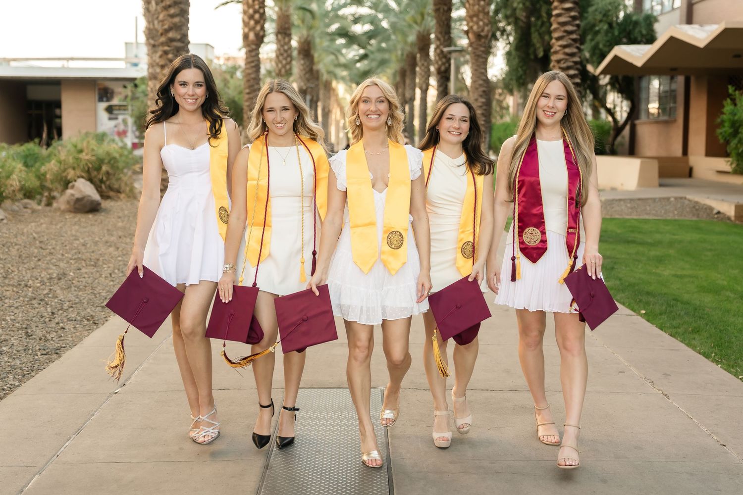 Group of graduates in maroon and yellow regalia walk together on campus path.