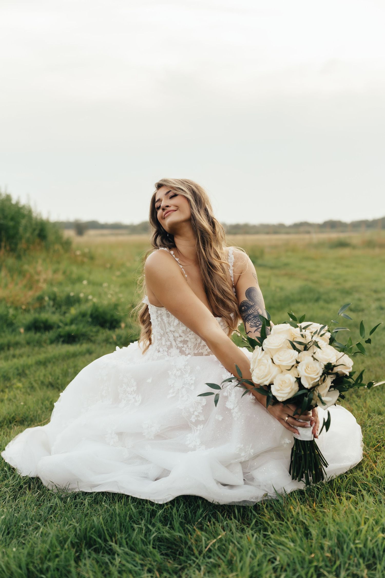 A romantic bridal portrait series showing someone in a flowing white wedding dress laying in green grass with white roses.