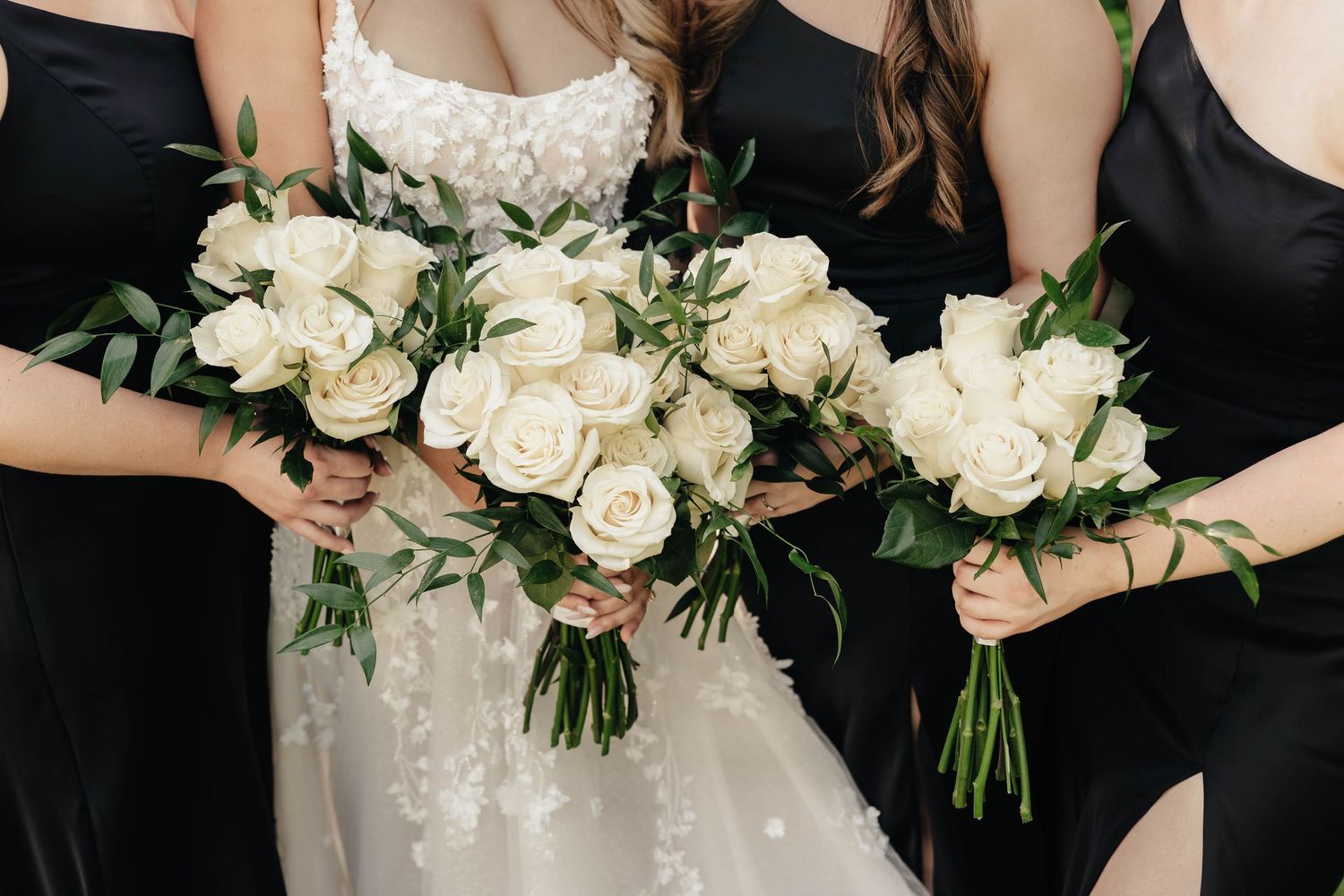 A row of wedding party members hold elegant white rose and greenery bouquets against black dresses.