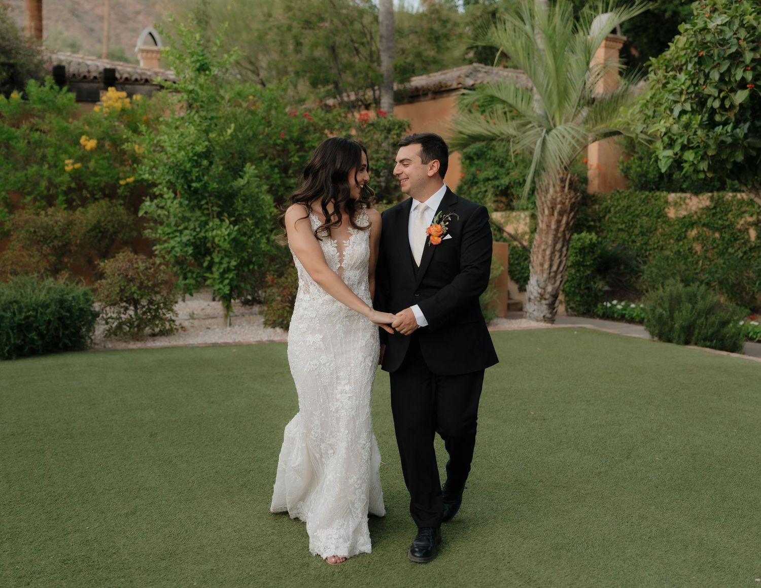 Bride in white lace gown and groom in black tuxedo holding hands on green lawn at outdoor wedding venue.