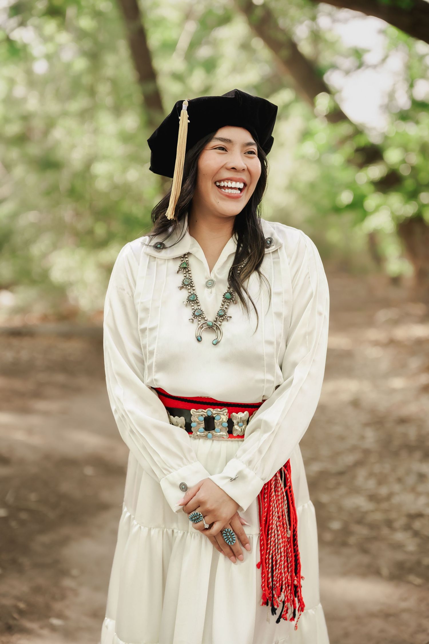 Graduate in traditional white dress with red sash and black cap in outdoor setting.